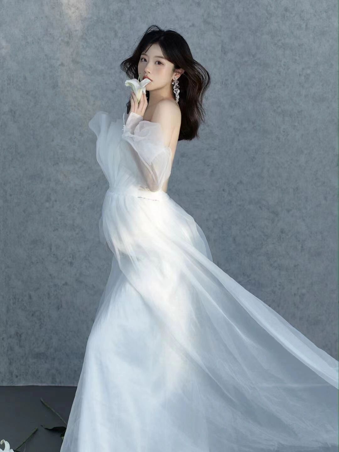 Woman in flowing white off-shoulder tulle gown holding white flower in elegant bridal portrait photography.