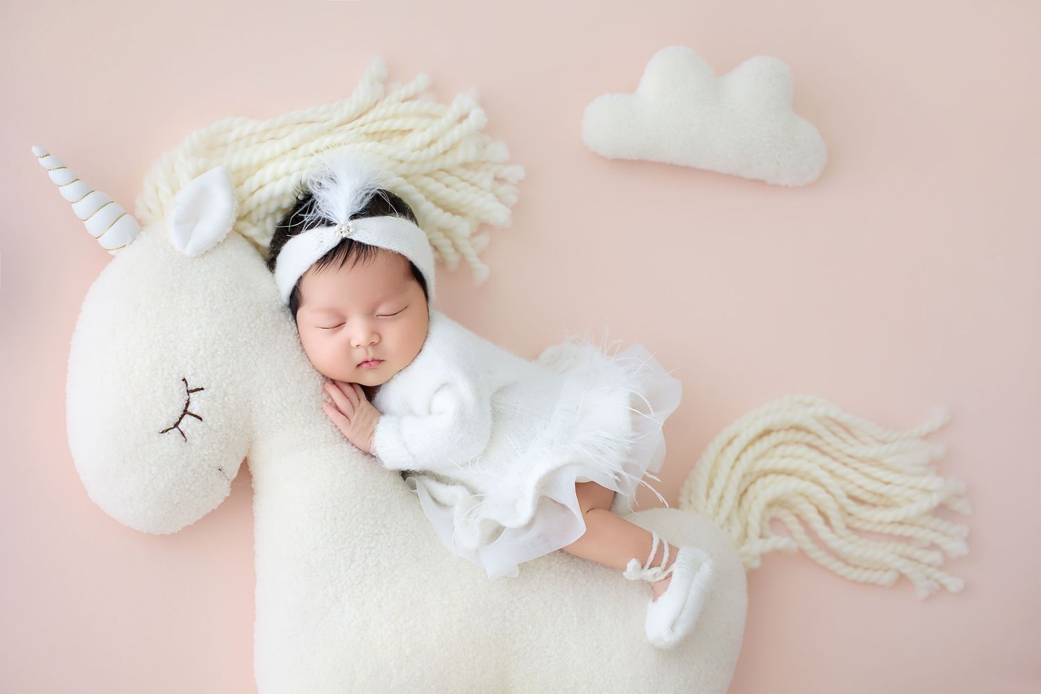 Peaceful newborn baby in white tutu dress resting on a unicorn plush prop with cloud decor on pink backdrop.