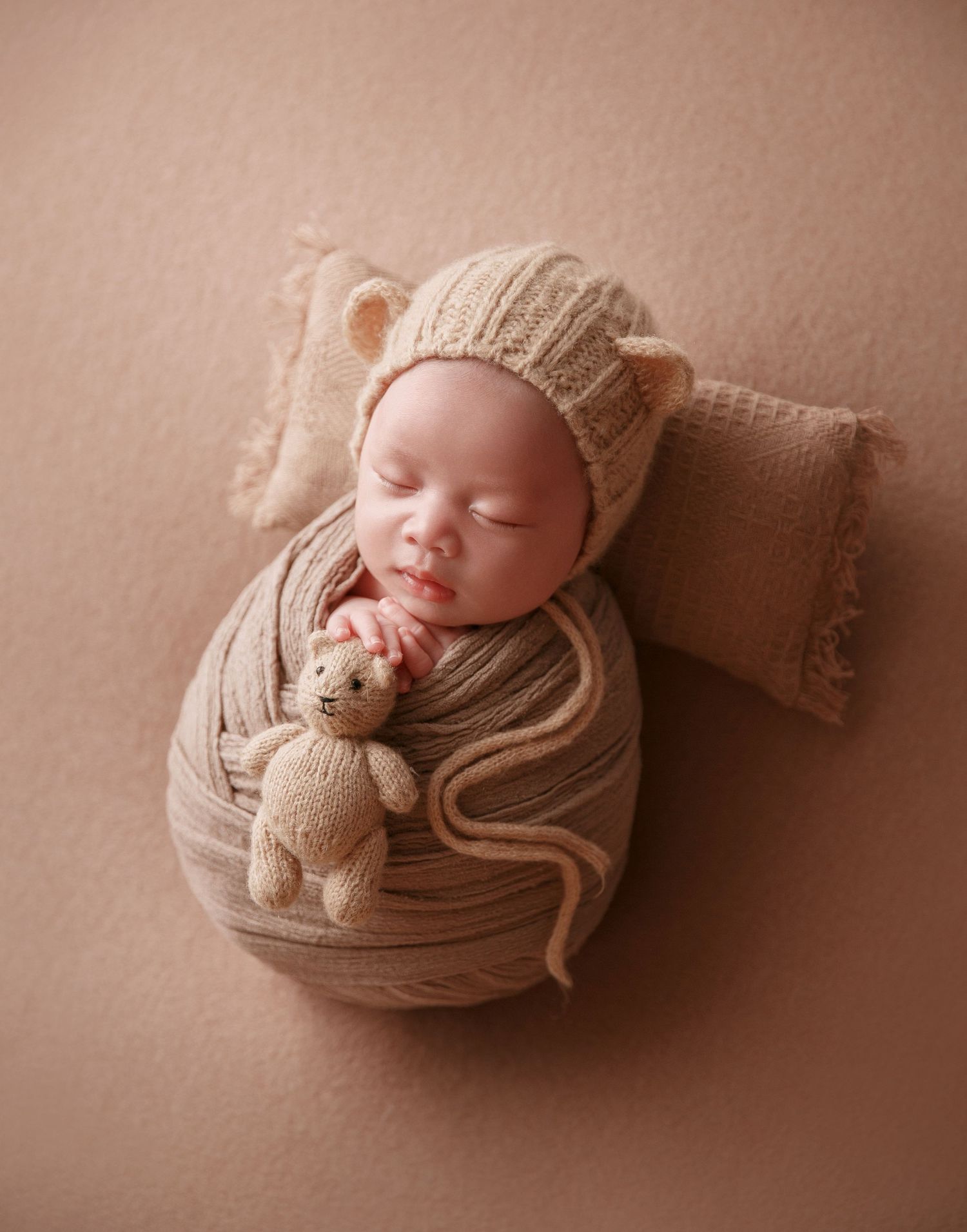 Newborn baby wrapped in beige knit blanket with bear ear bonnet holds small stuffed teddy bear.