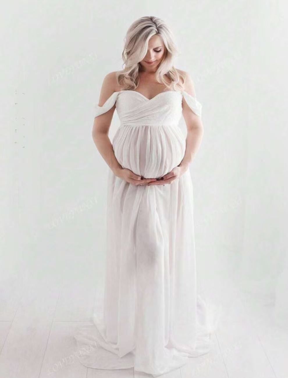 Pregnant woman in white off-shoulder draped chiffon maternity gown cradling baby bump in white studio.