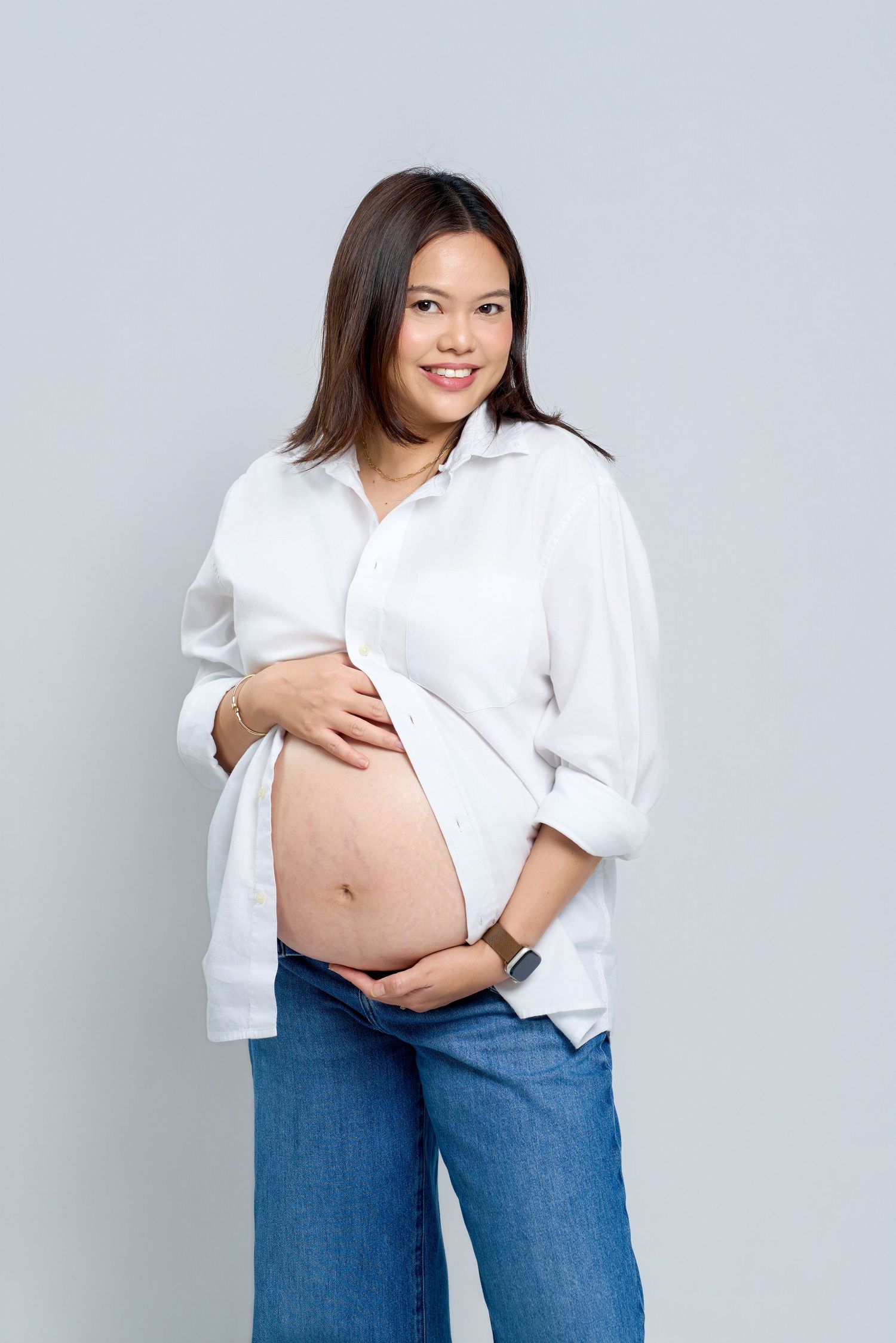 Smiling pregnant Asian woman in white shirt and jeans cradling her baby bump against a light gray background.