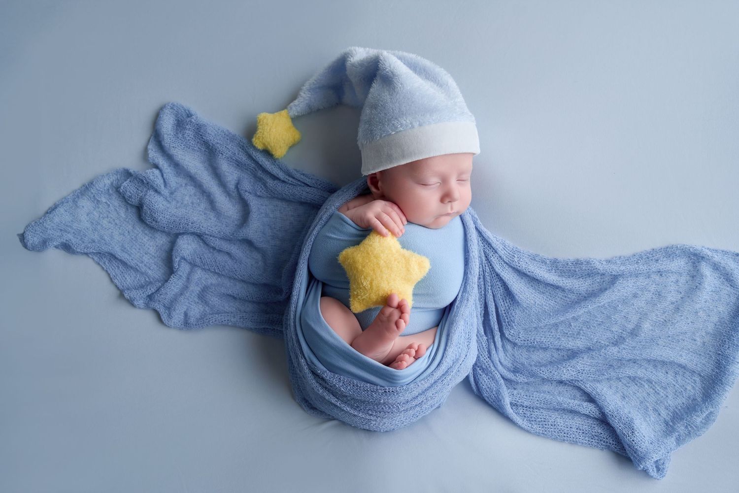 Sleeping newborn baby wrapped in blue knit blanket wearing star hat, holding yellow star props on blue background.