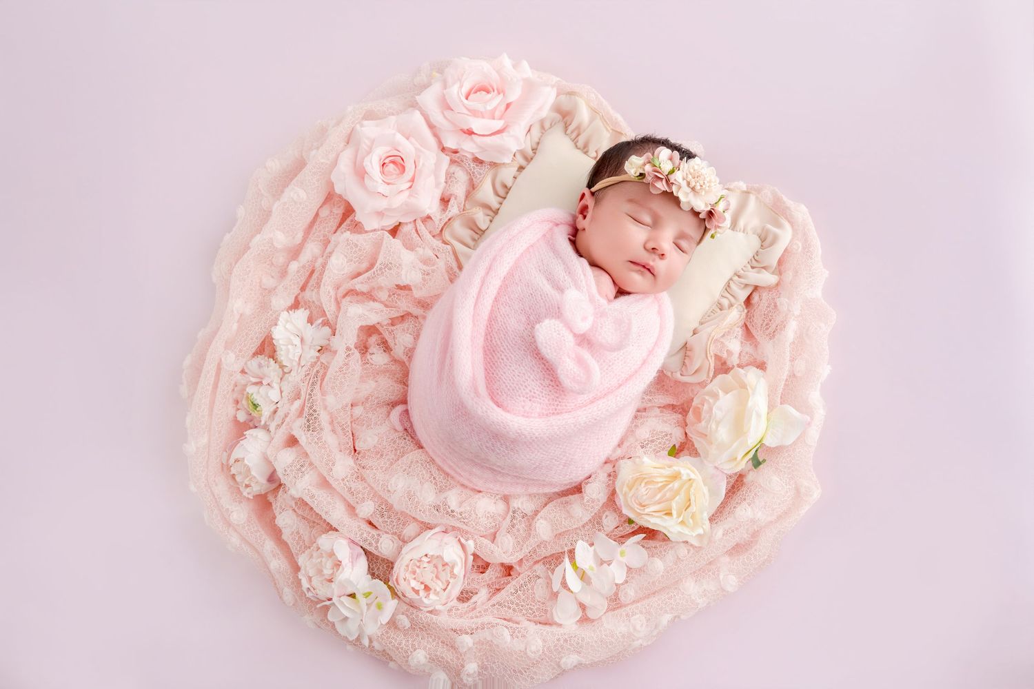 Newborn baby girl sleeping peacefully in pink floral wrap, nestled in a heart-shaped bed with roses.