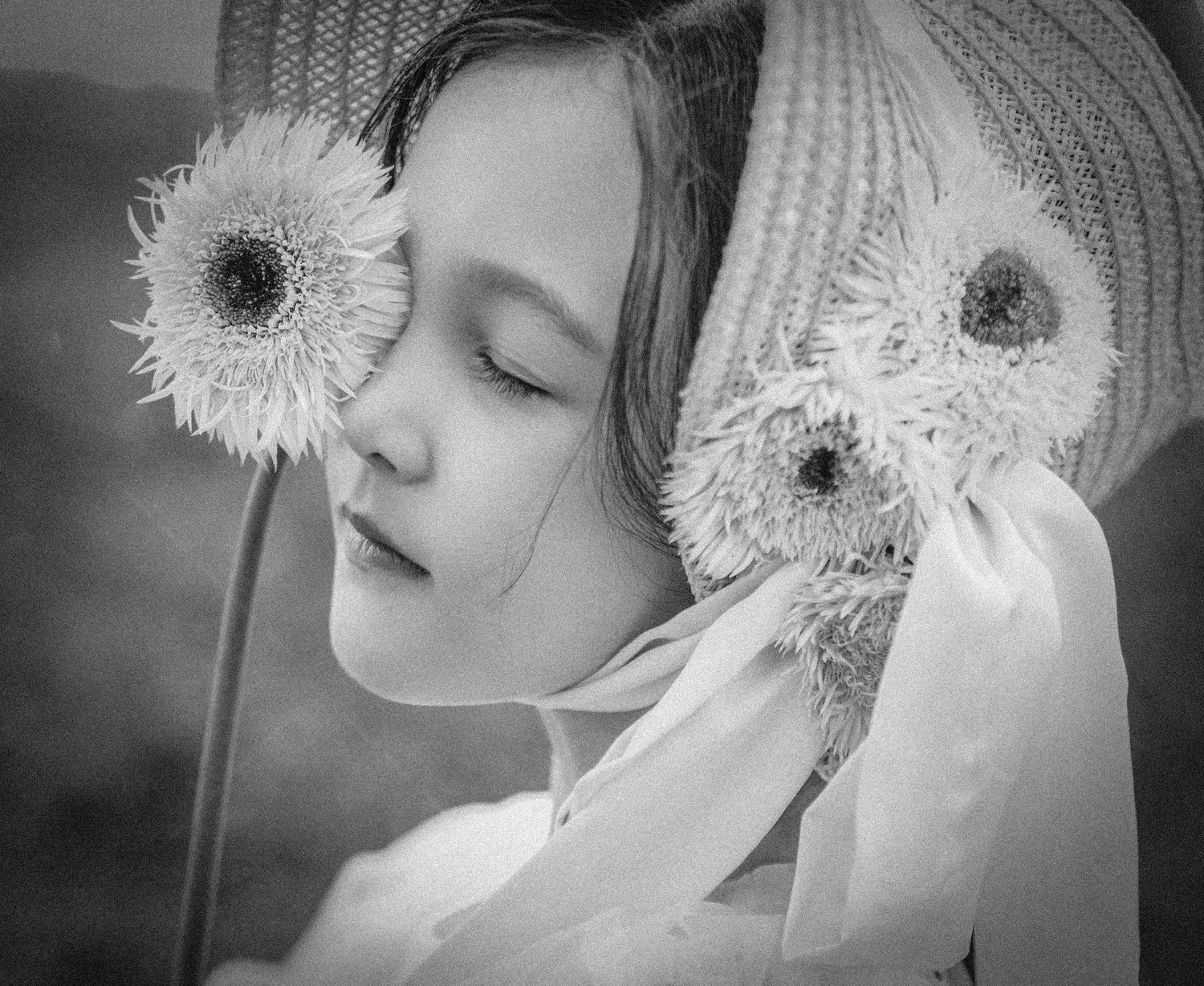Black and white artistic portrait of a young girl in straw hat holding large dandelion flowers near her face.