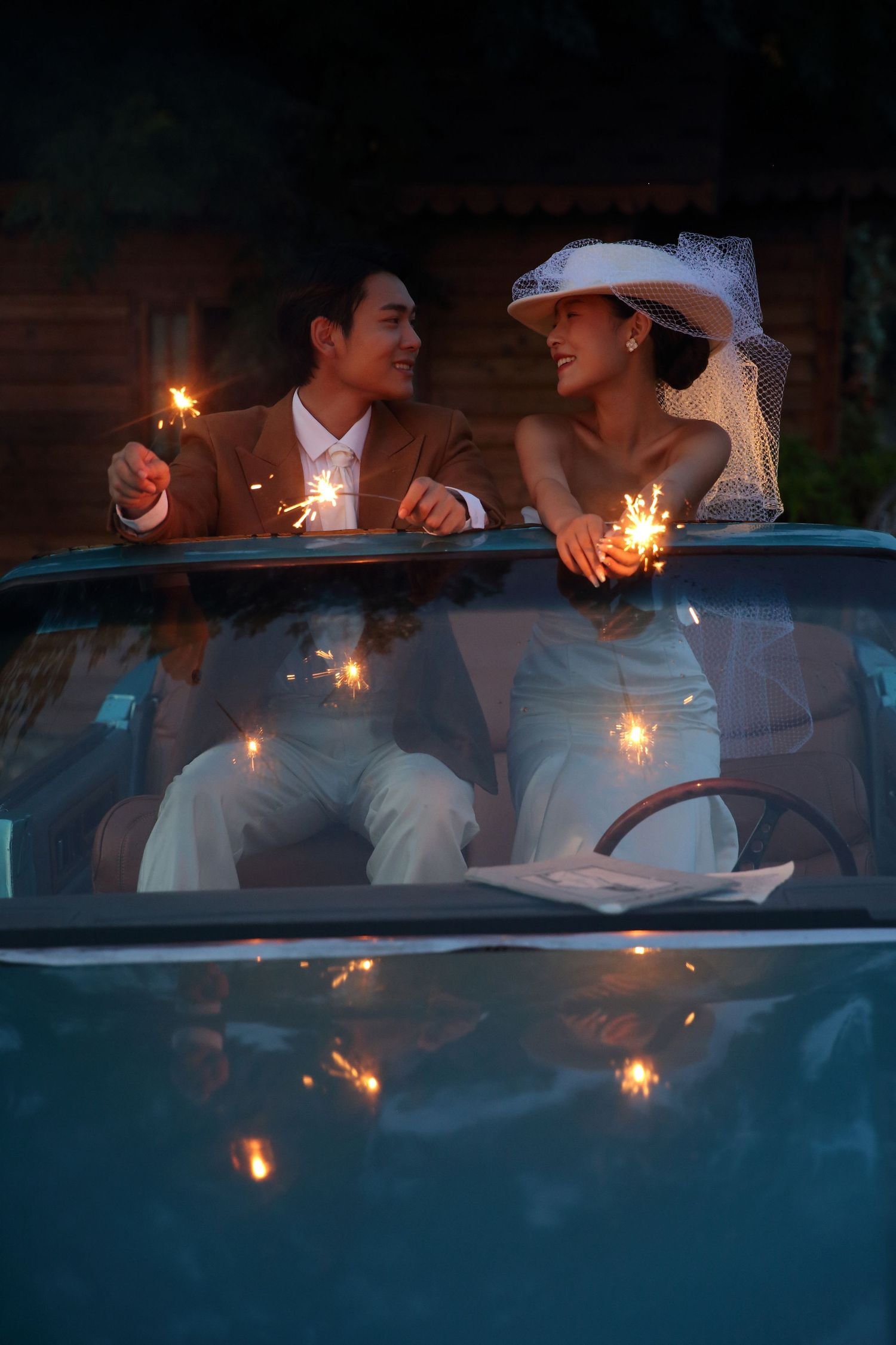 Bride and groom holding sparklers in a teal vintage convertible car at night, smiling joyfully.