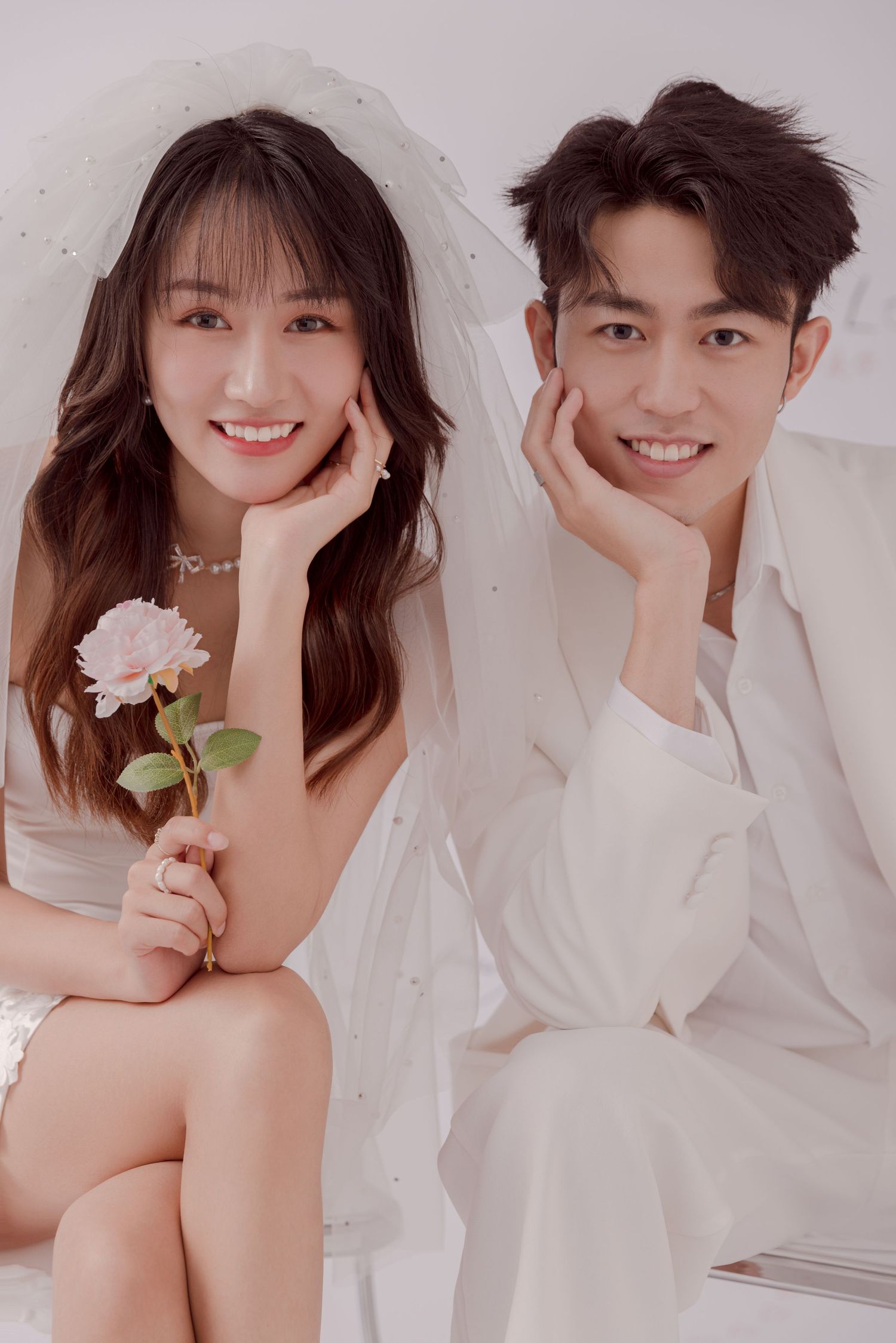 A smiling couple in white wedding attire sit together, the bride holding a pink rose under a sheer veil.