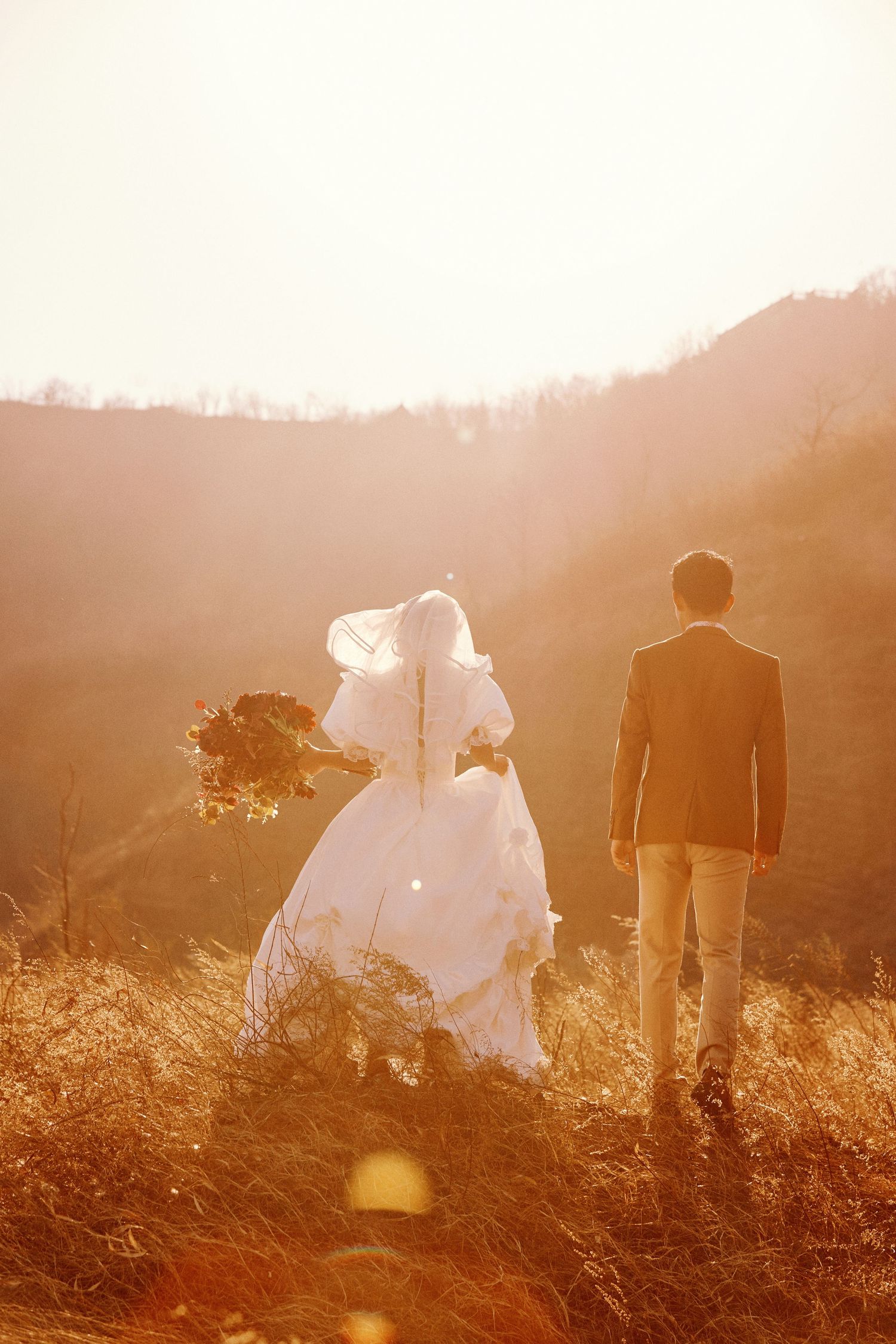 Bride and groom walk through golden sunlit fields with misty mountain backdrop in romantic wedding photo.