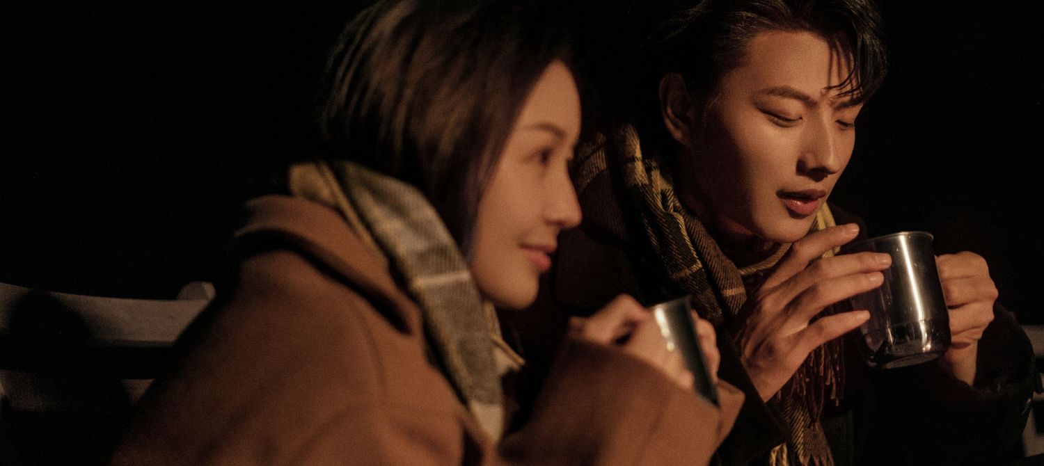 A young couple in warm coats sharing a cozy moment together while holding mugs in a dimly lit setting.