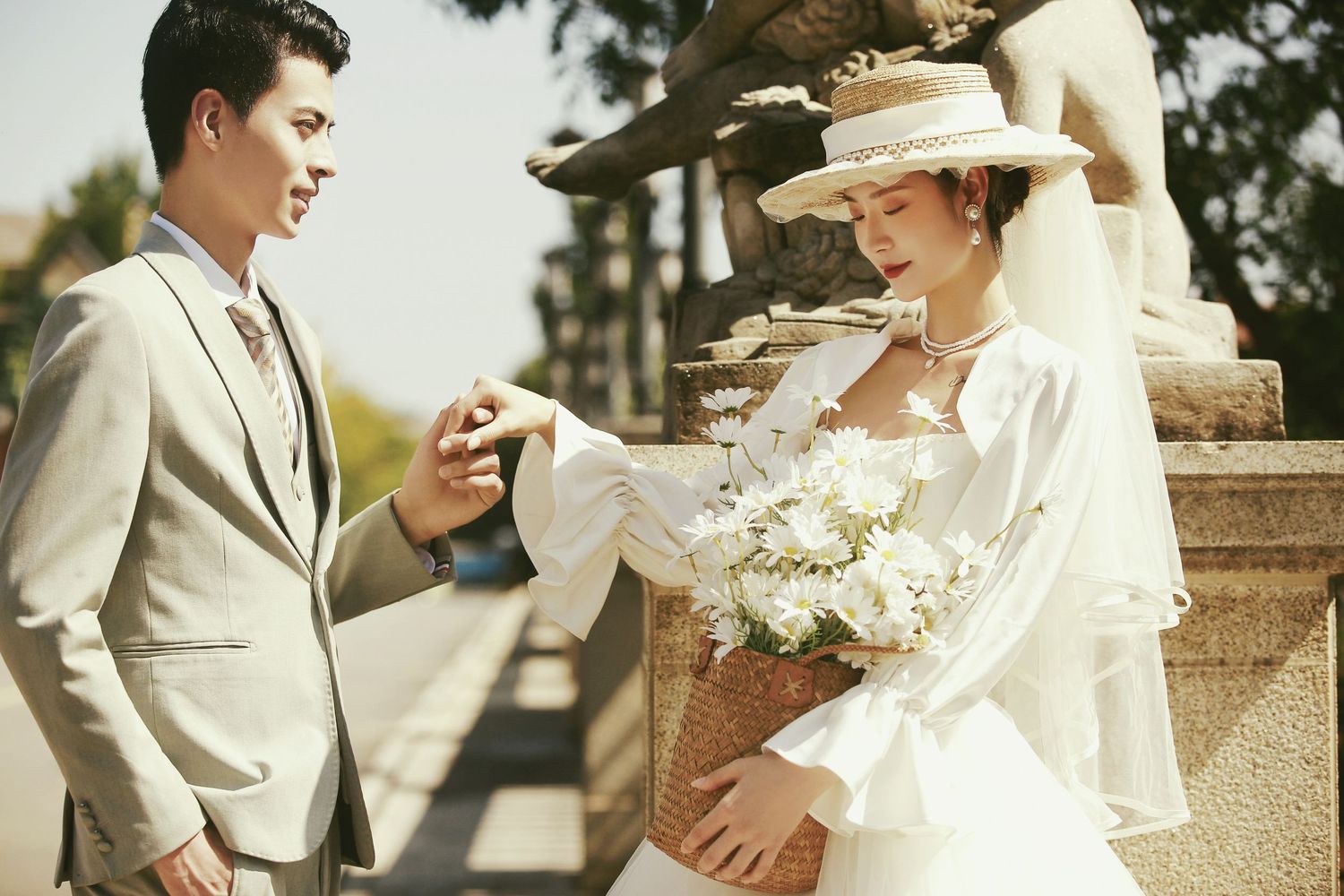 Groom tenderly holds bride's hand near classical stone sculpture, bride wearing white gown and straw hat.