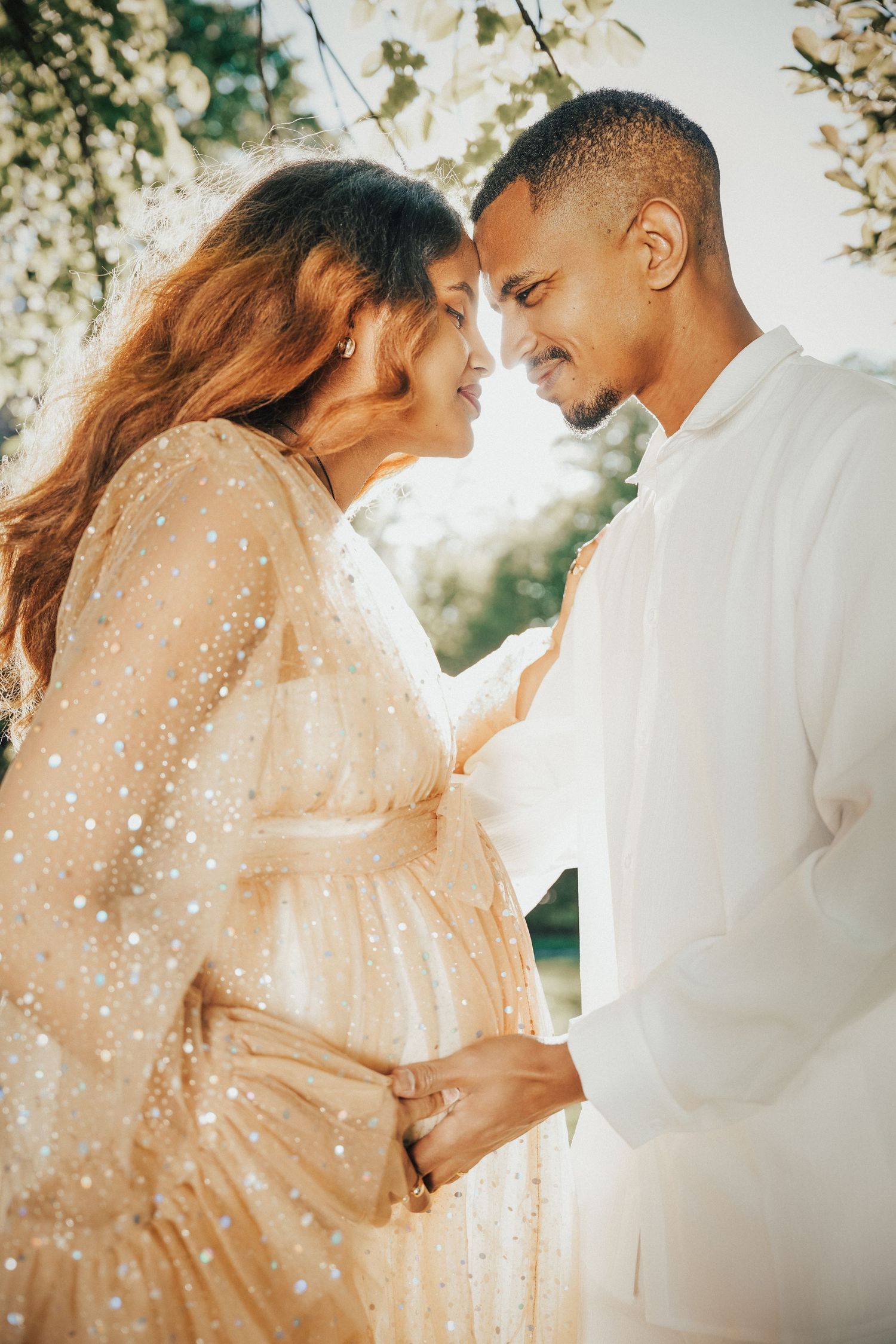 A pregnant couple in elegant attire shares a tender moment outdoors among blooming trees in warm golden light.