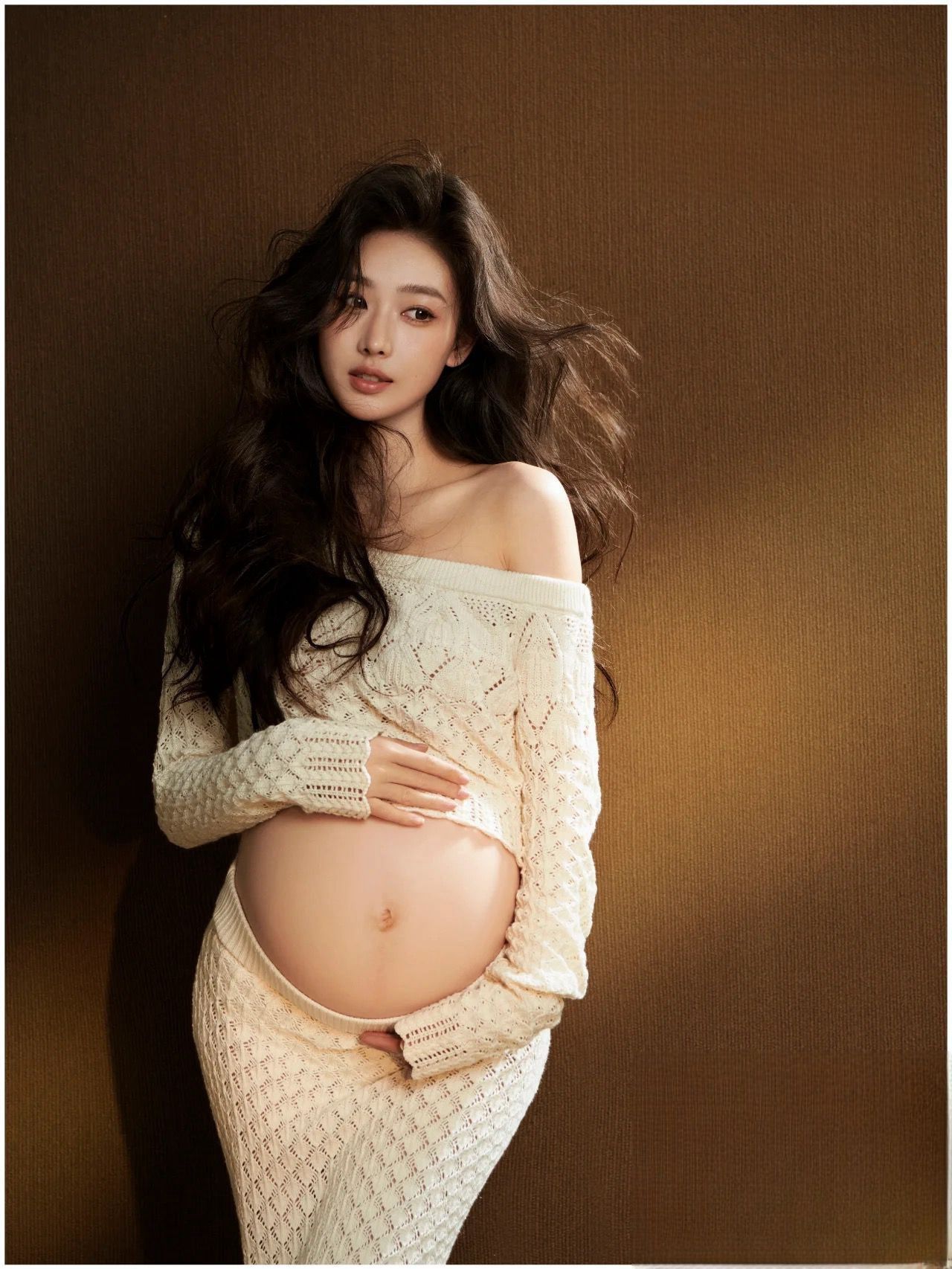Elegant pregnant model with long dark hair in cream off-shoulder cable-knit maternity outfit.
