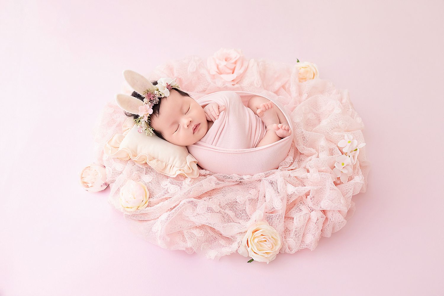 Newborn baby girl wrapped in pink lace swaddle with bunny headband surrounded by cream roses.