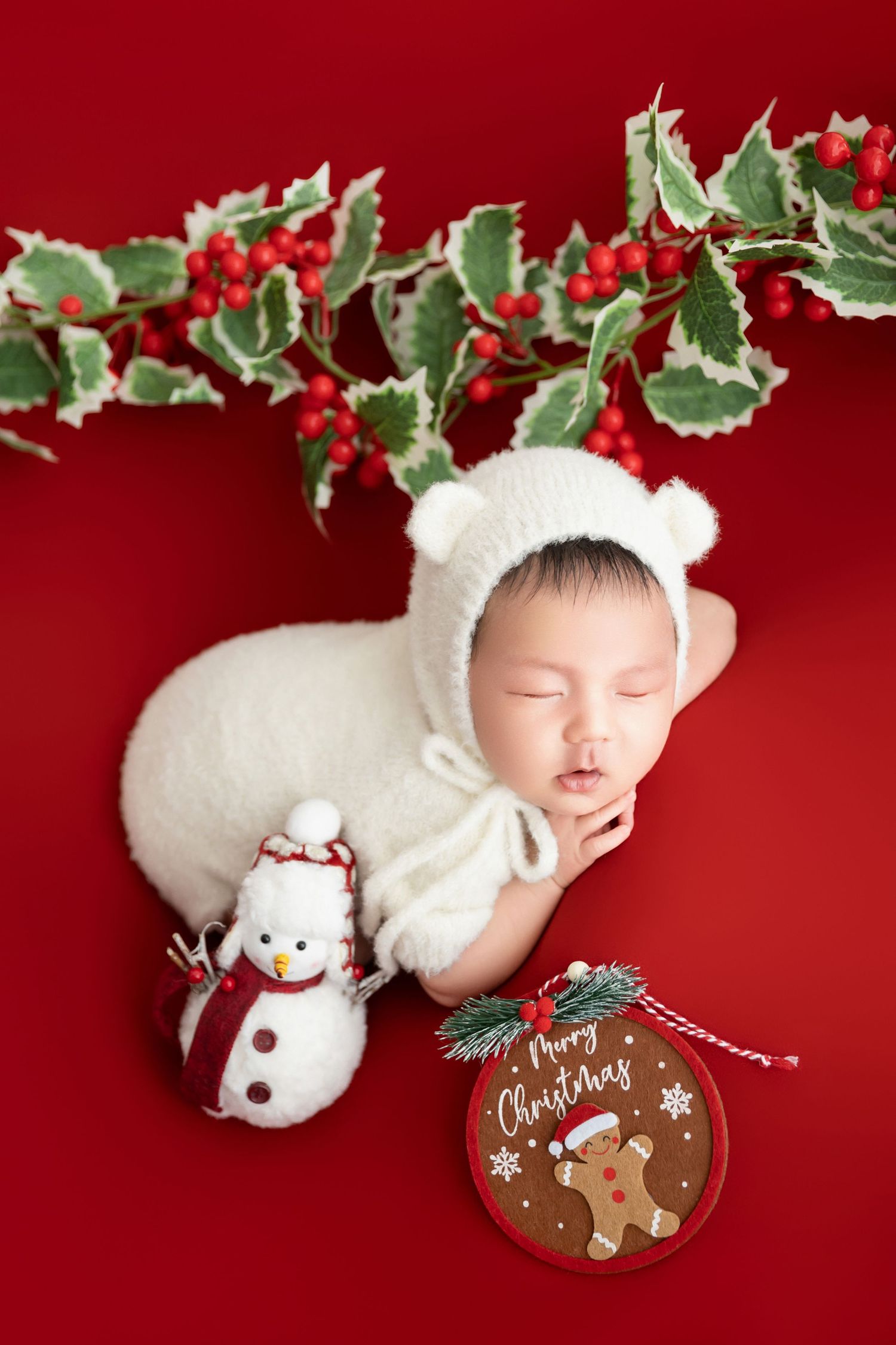 Peaceful newborn in white bear outfit rests on red background with snowman toy and Merry Christmas sign.
