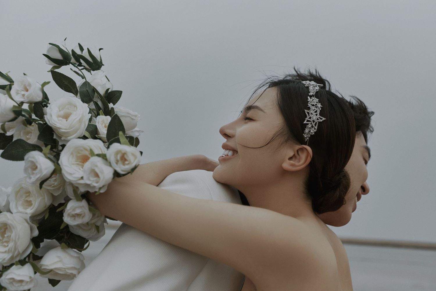 Smiling bride in white gown embraces groom while holding bouquet of white roses on overcast day.