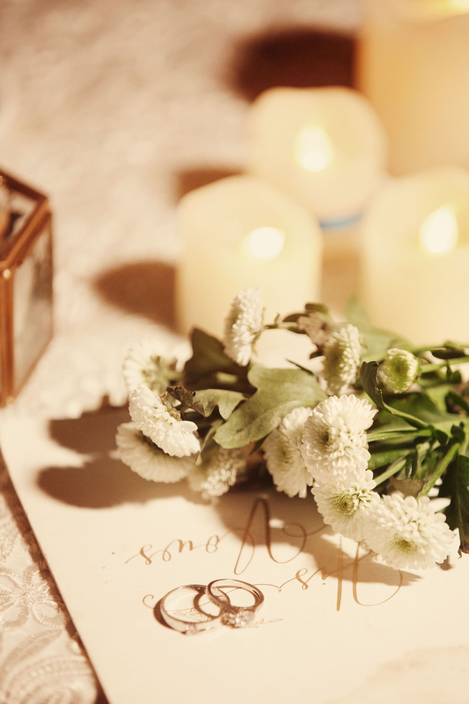 Wedding rings rest on white chrysanthemums beside glowing candles on a lace-covered table.