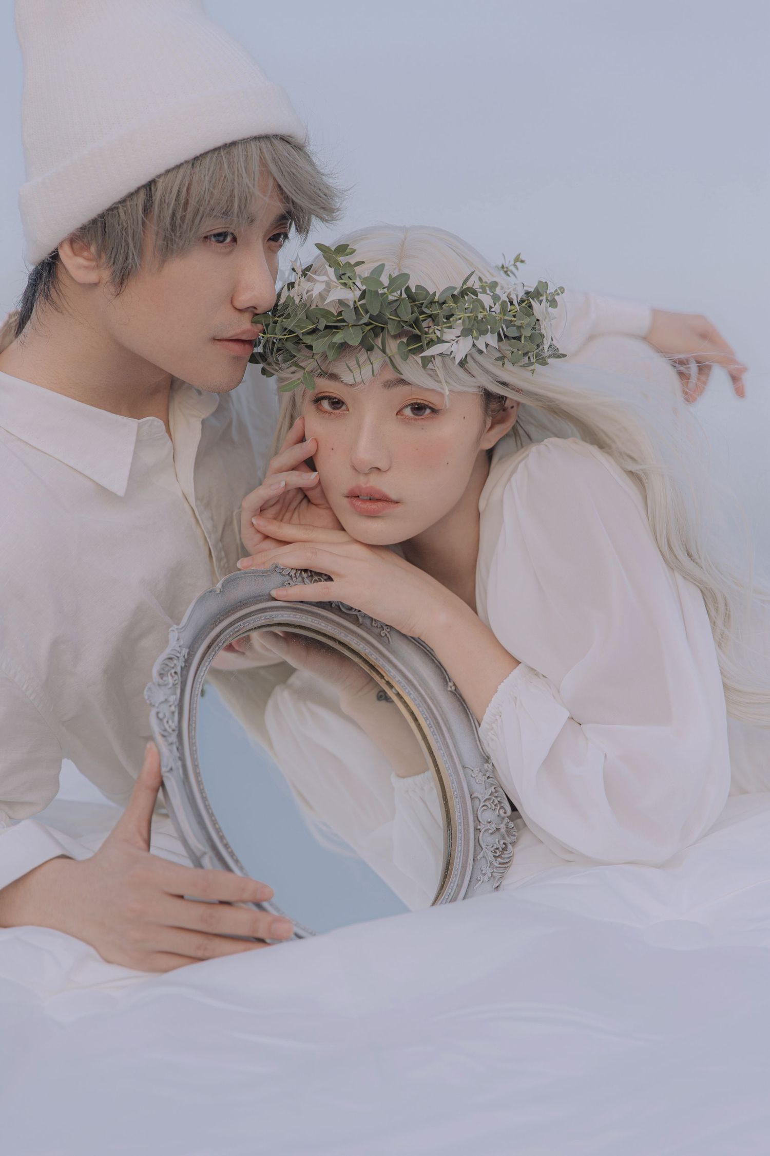 Couple in white clothing poses with oval mirror, woman wearing green leaf crown resting chin on hands.