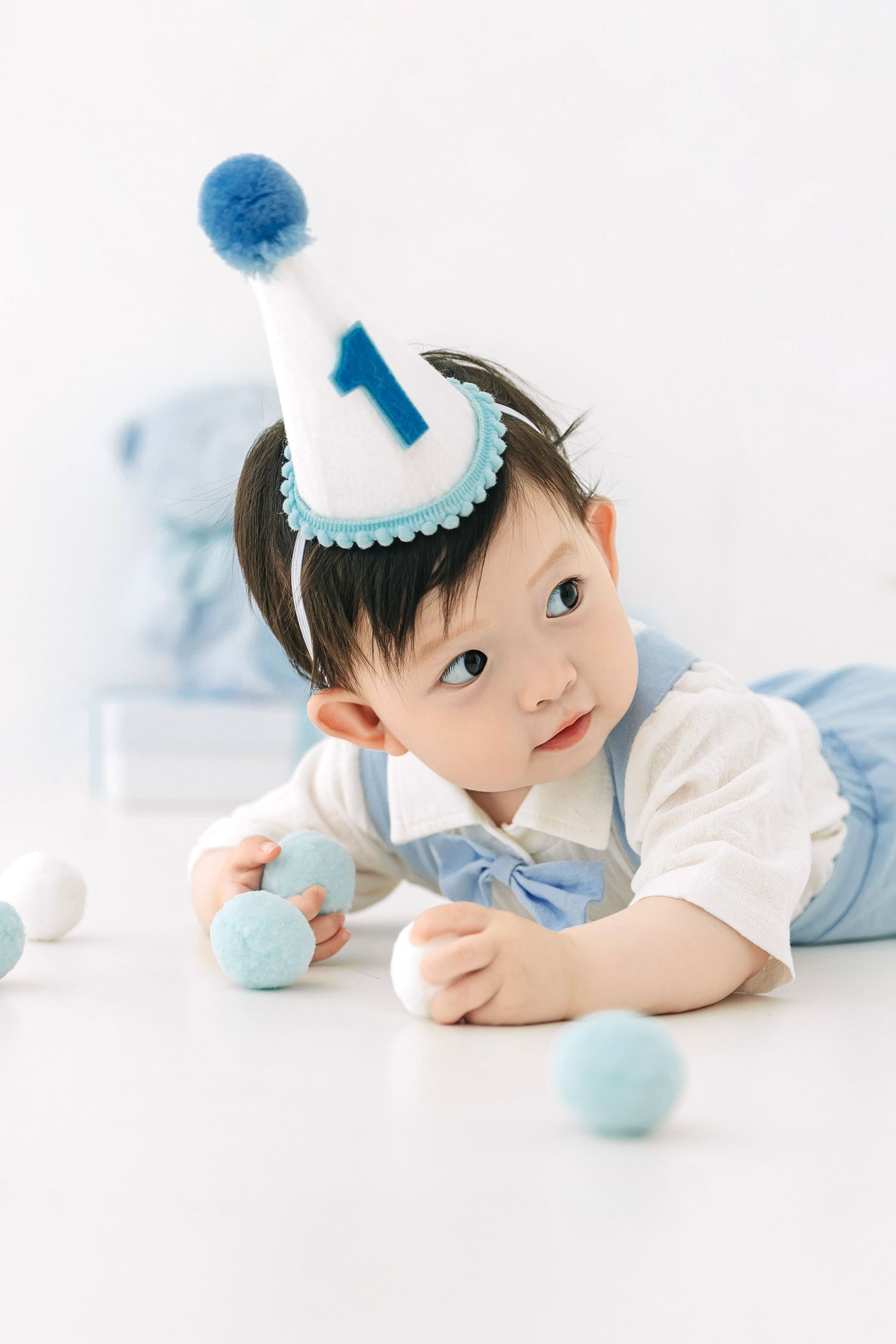 Cute baby boy in blue outfit and number 1 birthday hat playing with colorful pom poms on white floor.