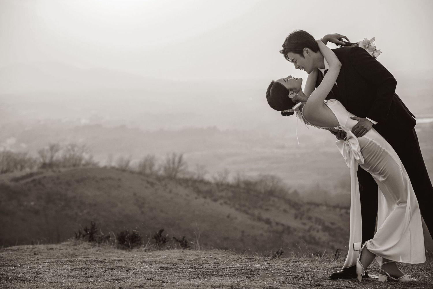 Black and white romantic wedding photo of groom dipping bride against a scenic hilltop landscape backdrop.