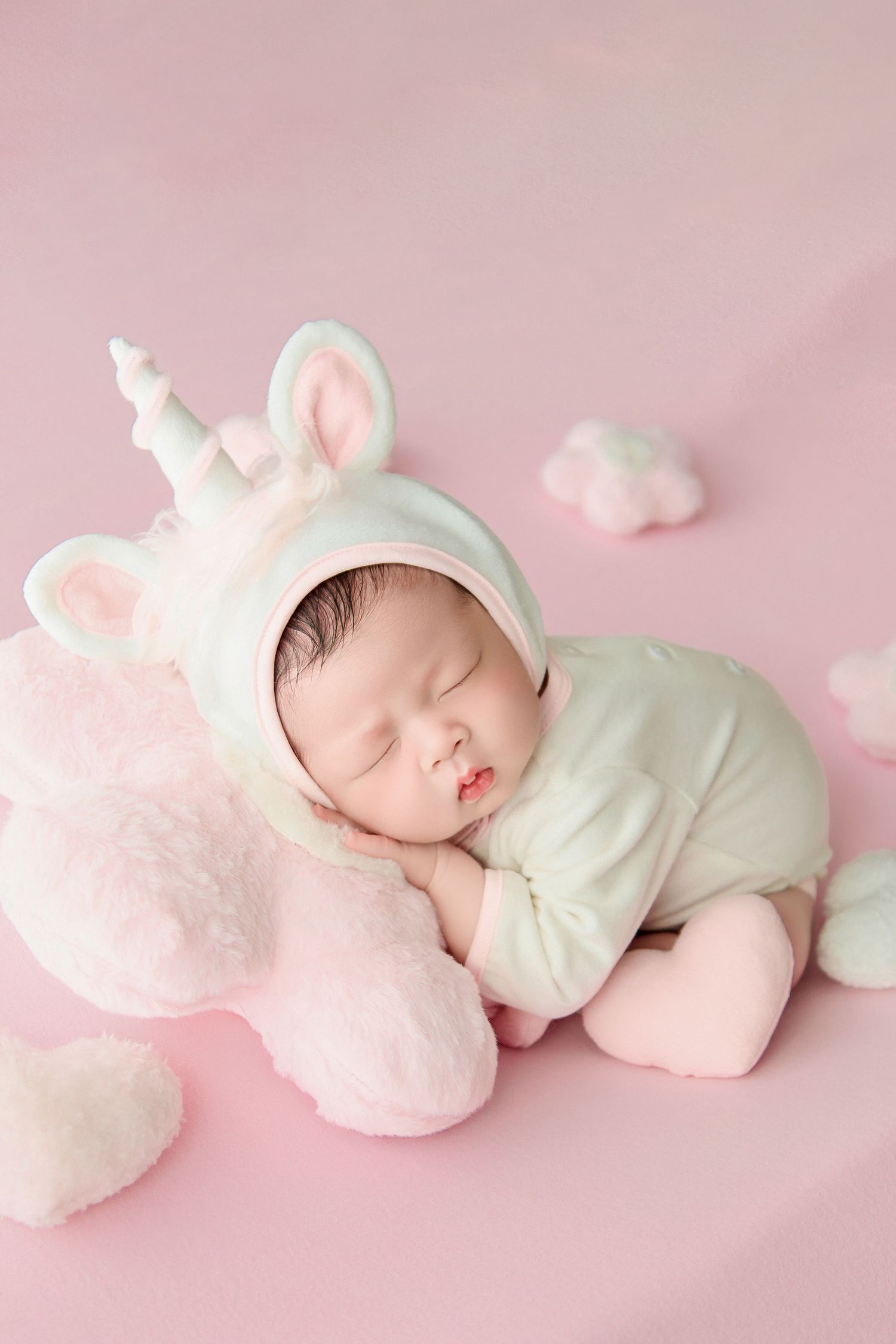 Sleeping newborn baby in white unicorn costume on pink background with fluffy clouds and stuffed unicorn toy.