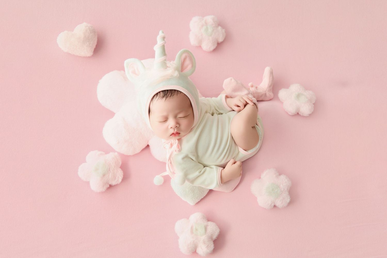 Sleeping newborn baby in white unicorn outfit and hat on pink background with fluffy cloud and flower decorations.