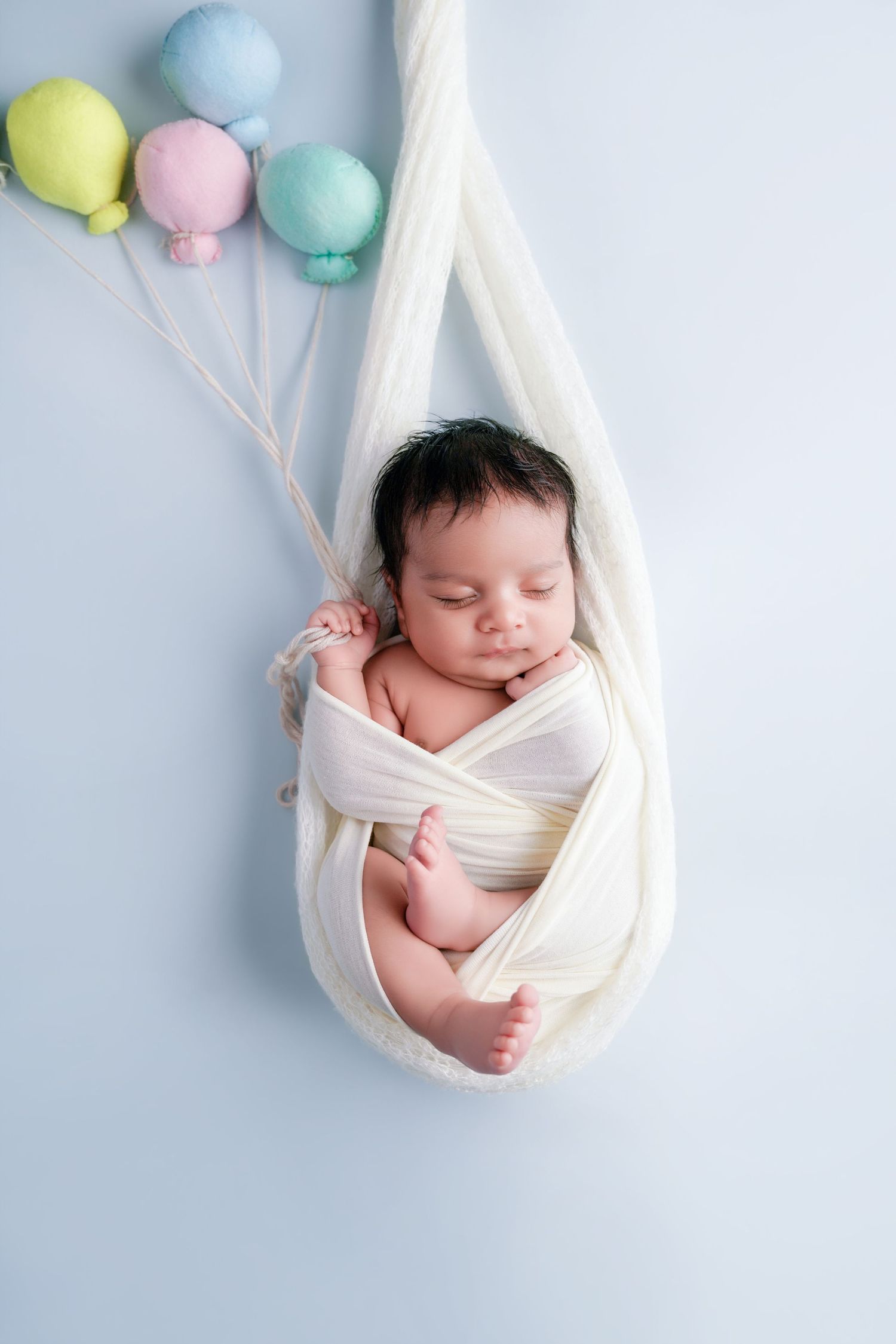 Newborn baby sleeping peacefully wrapped in white swaddle cloth, suspended with pastel balloon decorations on blue background.