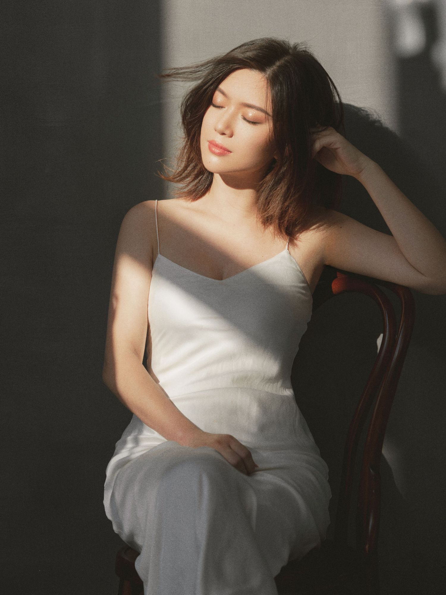 Young woman in white slip dress sits on chair with eyes closed, bathed in warm sunlight against a gray wall.
