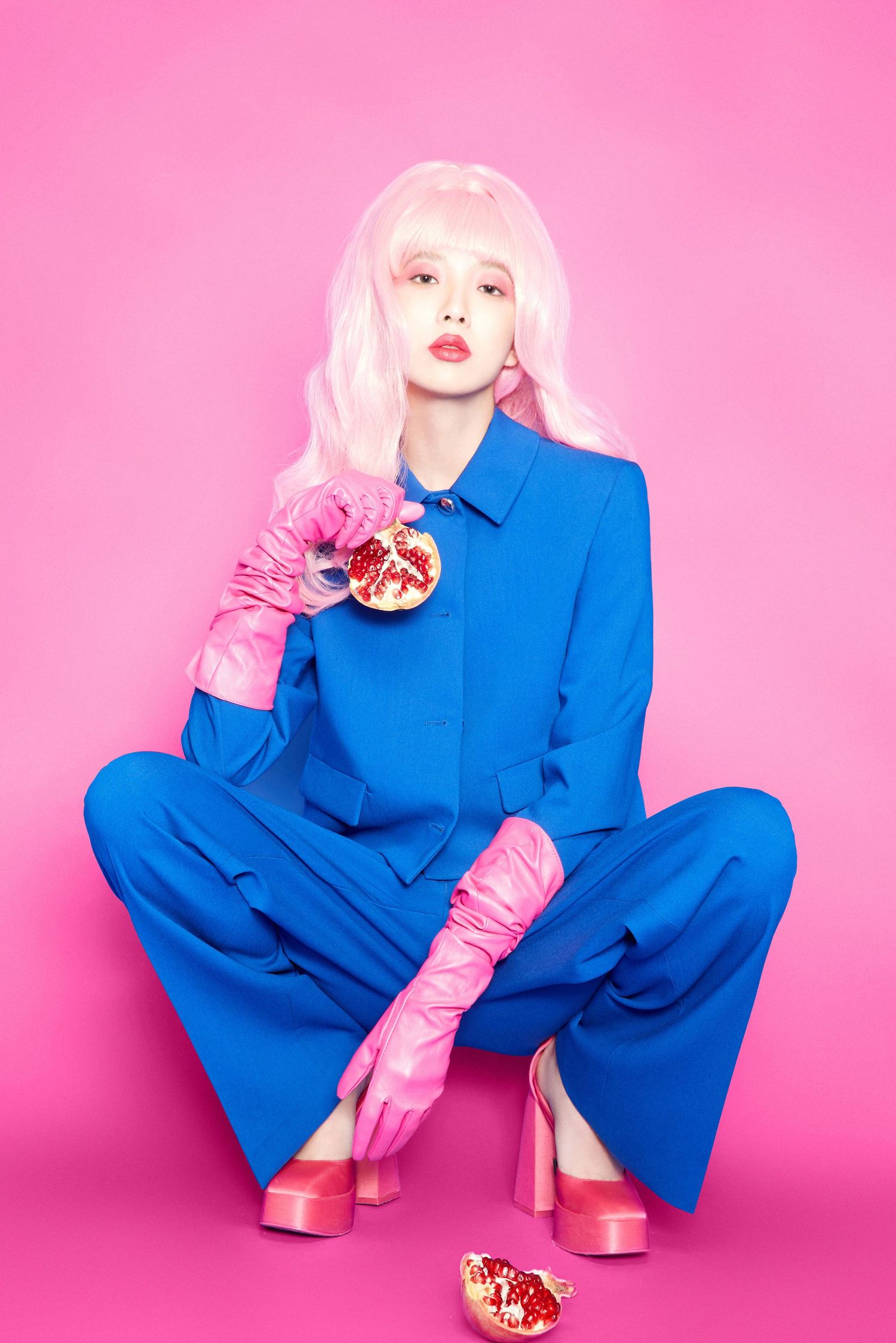 Model in blue suit with pink hair and gloves holds pomegranate on pink background.