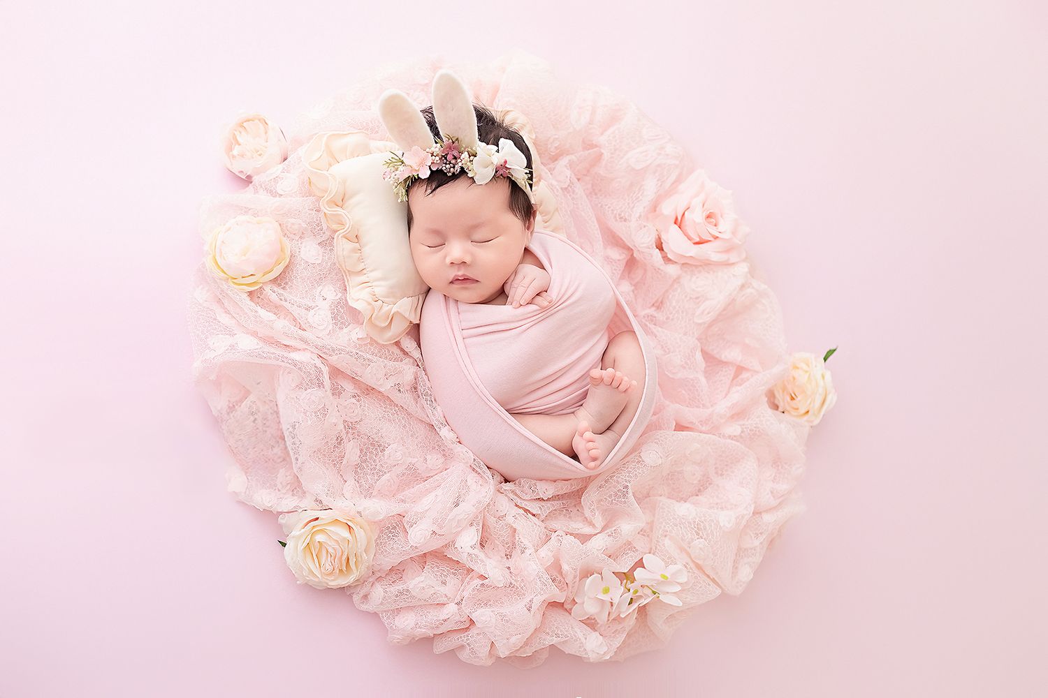 Sleeping newborn baby girl wrapped in pink swaddle with bunny ear headband in floral nest.