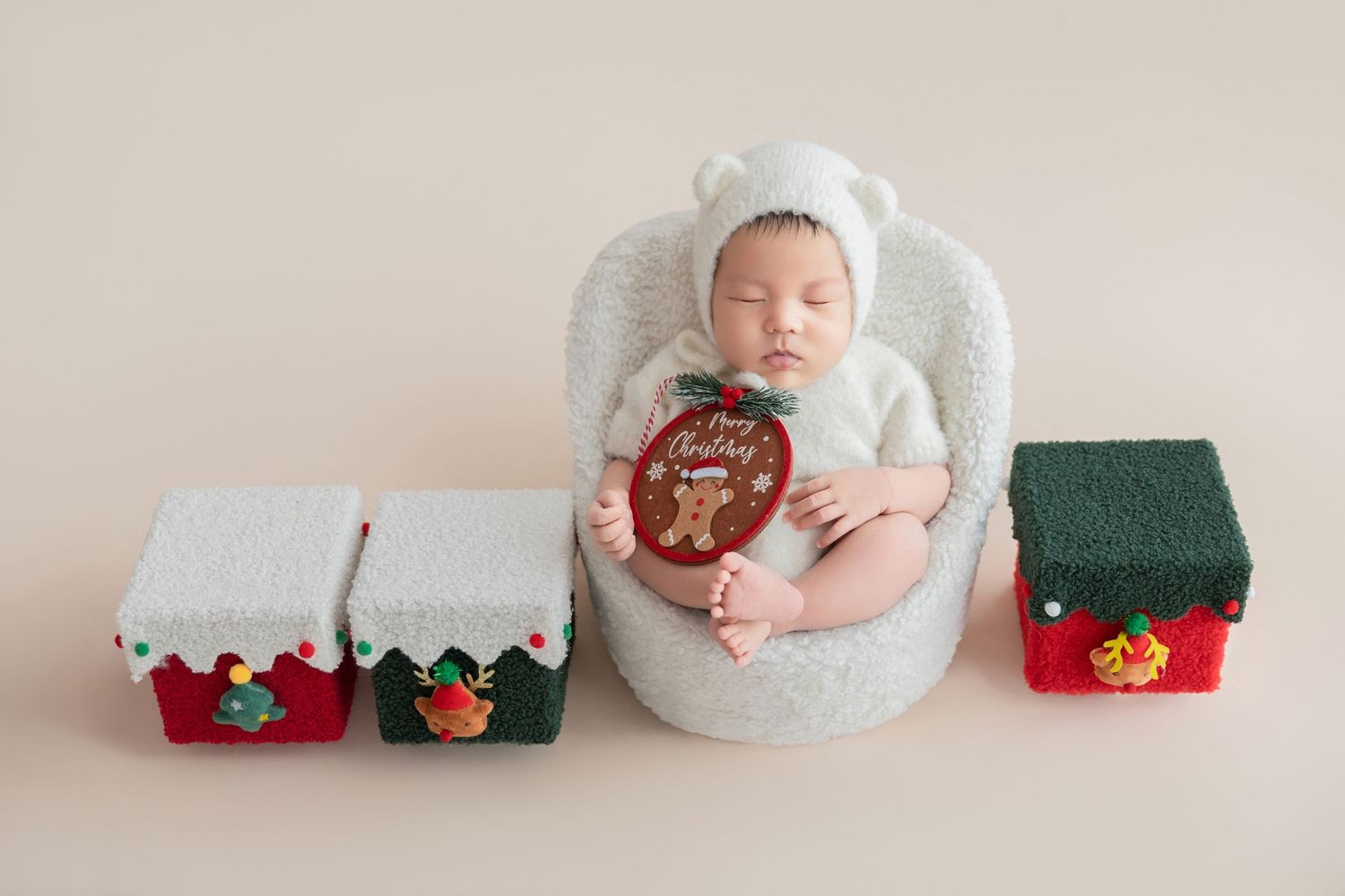 Sleeping newborn in white bear hat holds Merry Christmas ornament in tiny chair flanked by festive gift boxes.