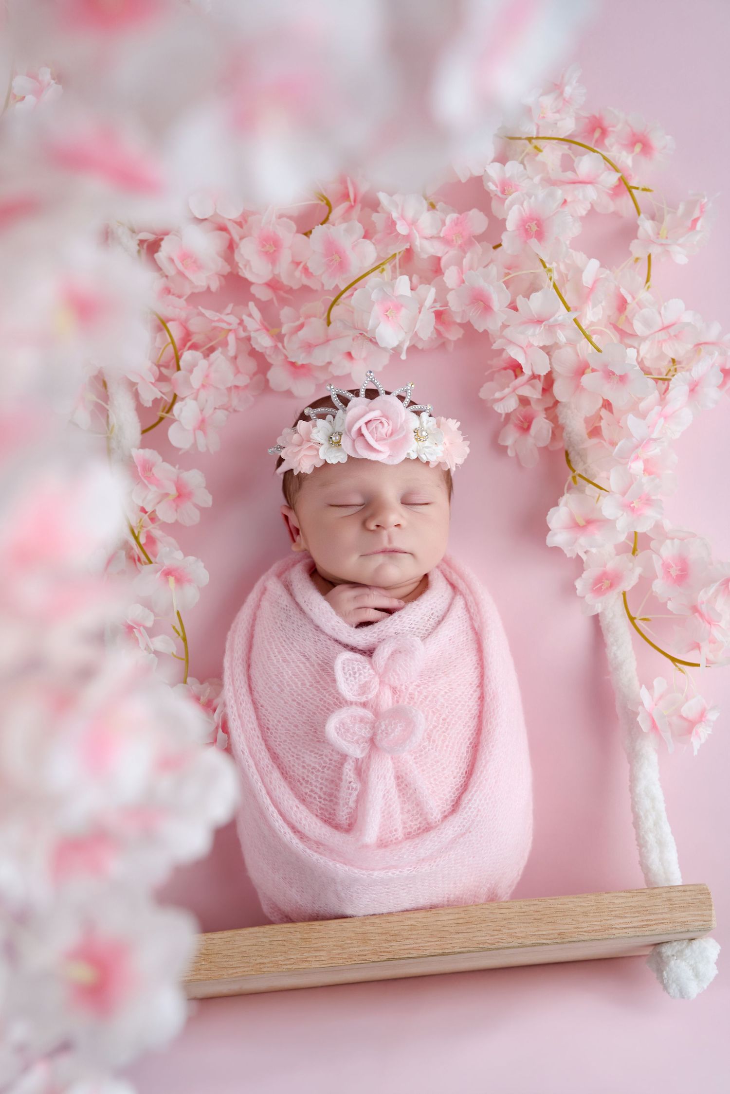 Newborn baby girl swaddled in pink wrap with floral headband sleeping on a swing decorated with pink cherry blossoms.