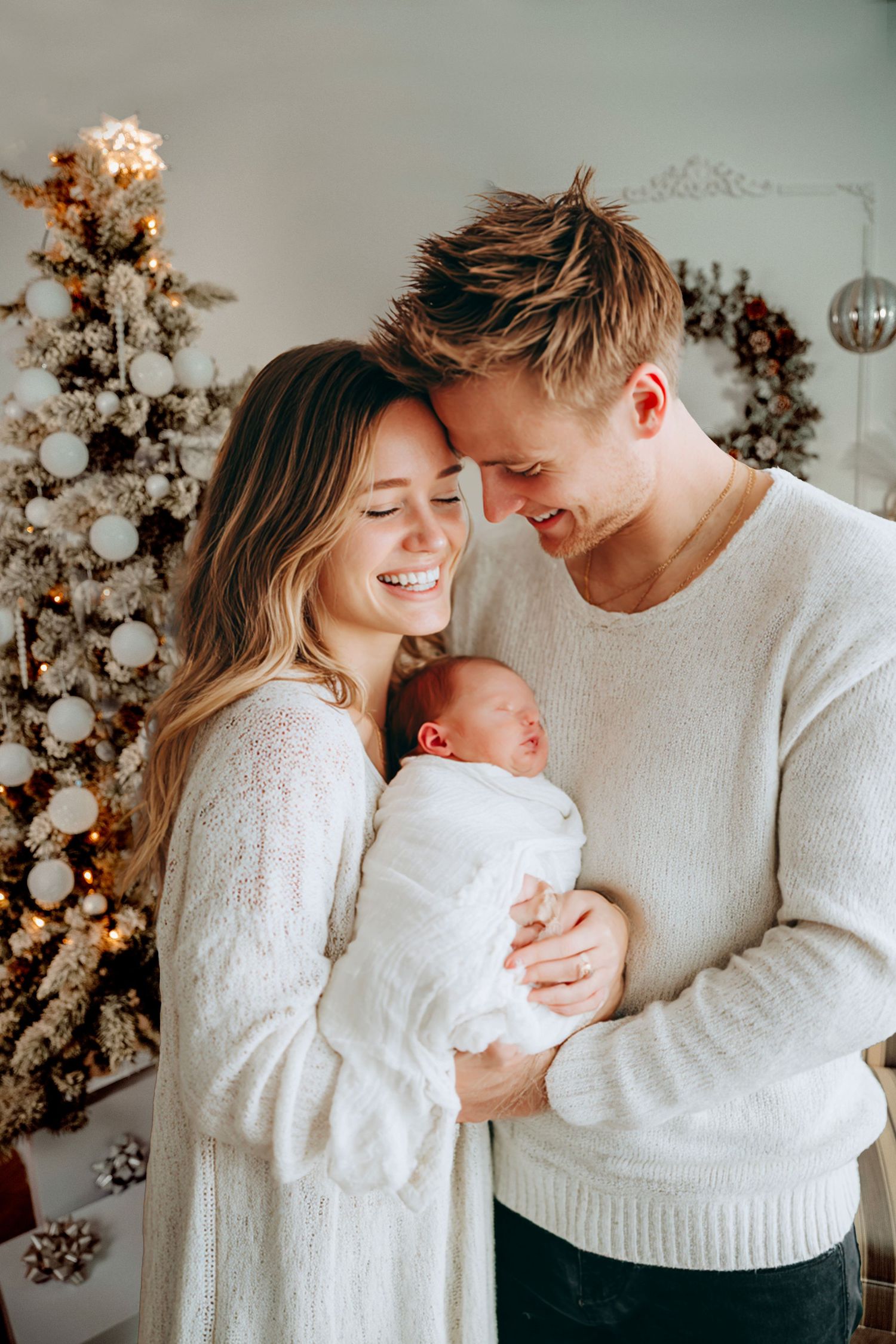 Happy young couple in white sweaters holding newborn baby in front of decorated Christmas tree.