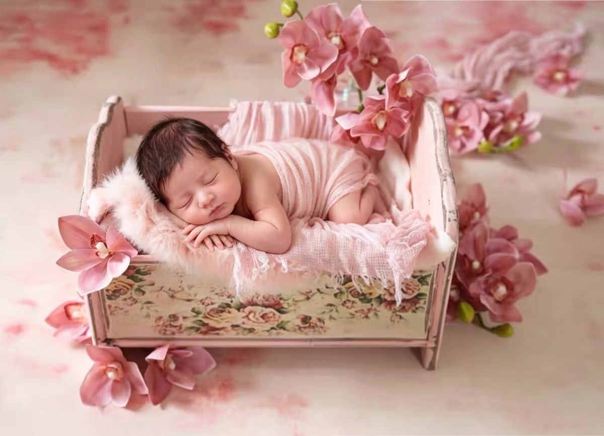 Newborn baby sleeping peacefully in a small vintage pink floral wooden bed surrounded by pink flowers.