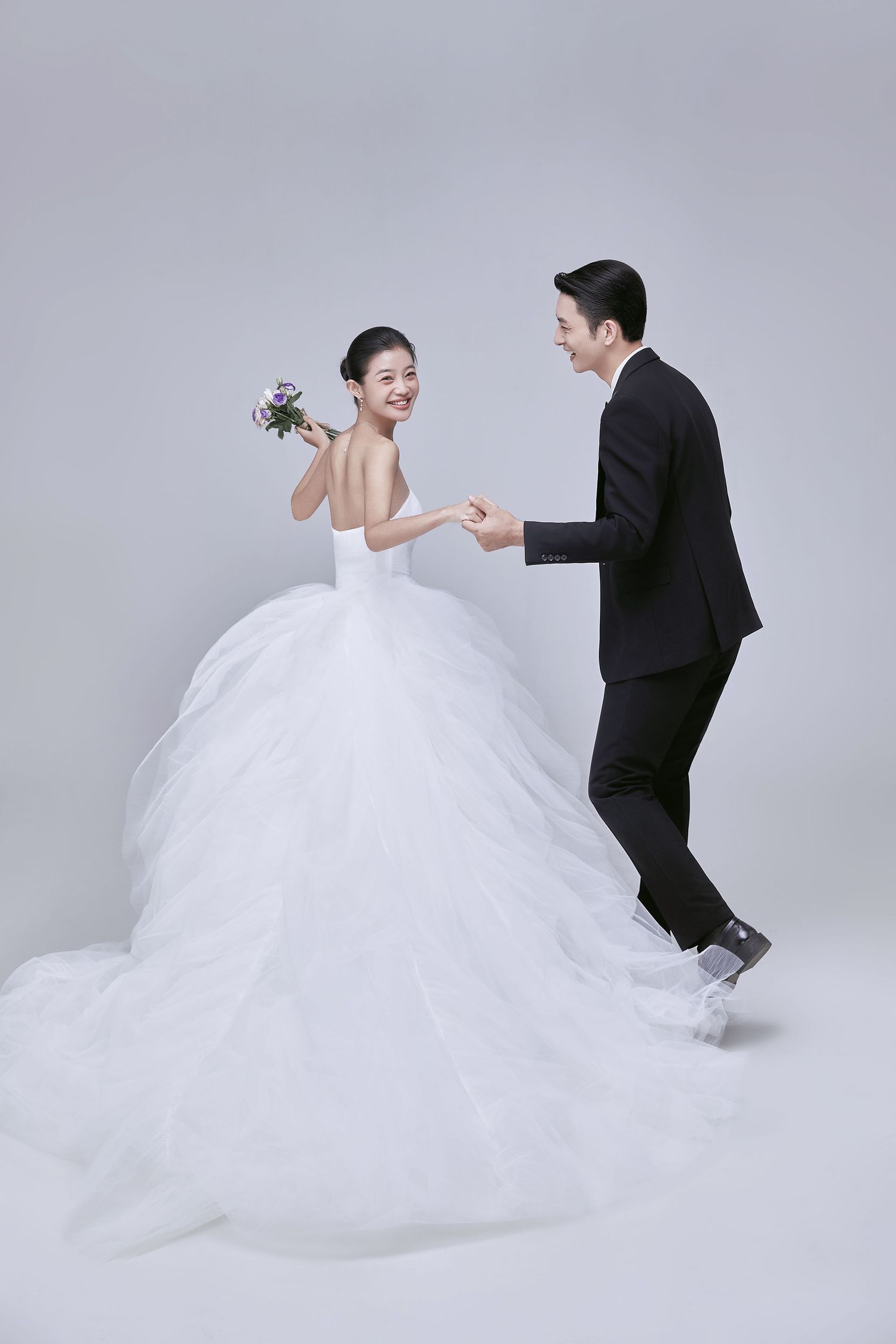 Happy Asian couple in wedding attire dancing together, bride holding bouquet in white ballgown.