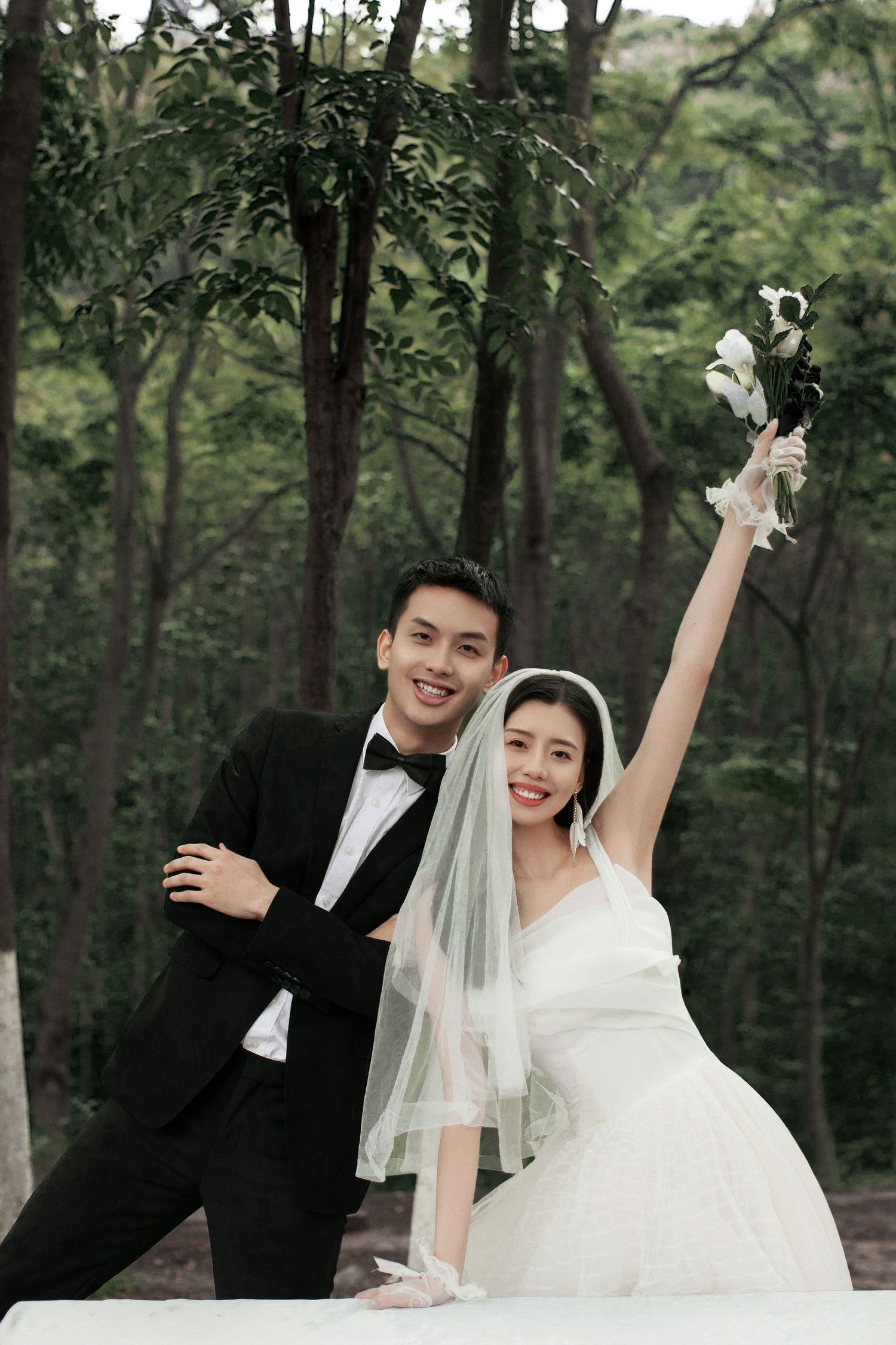Joyful bride raises bouquet triumphantly while posing with smiling groom in a lush green forest wedding portrait.