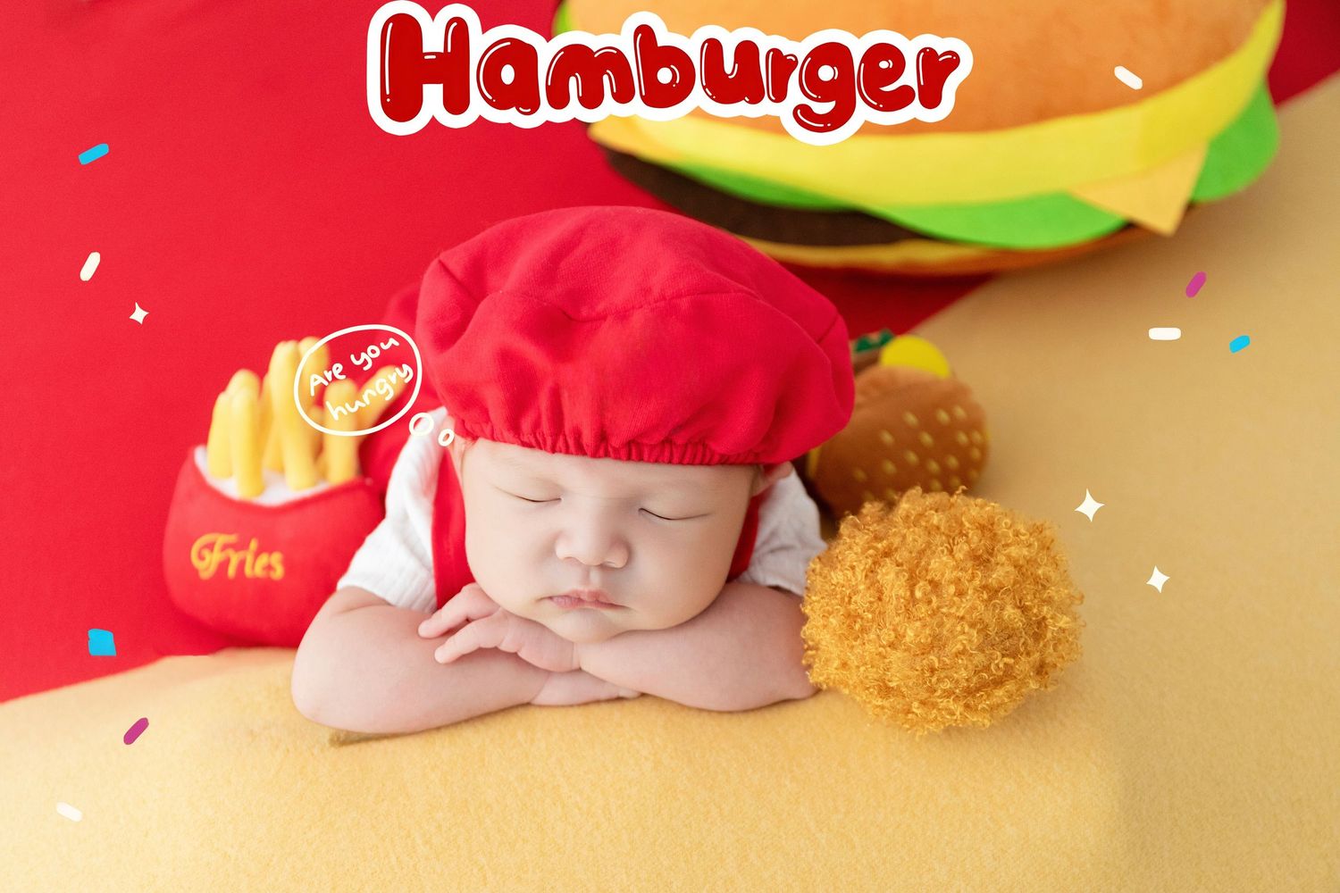 Sleeping baby dressed in red chef hat posed with toy fries and hamburger props in a fast food themed photo.