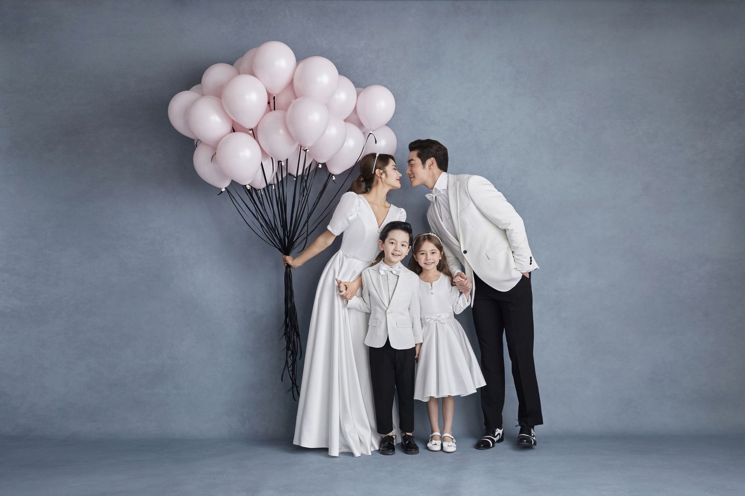 Family of four in white outfits posing with pink balloons in a romantic studio portrait.