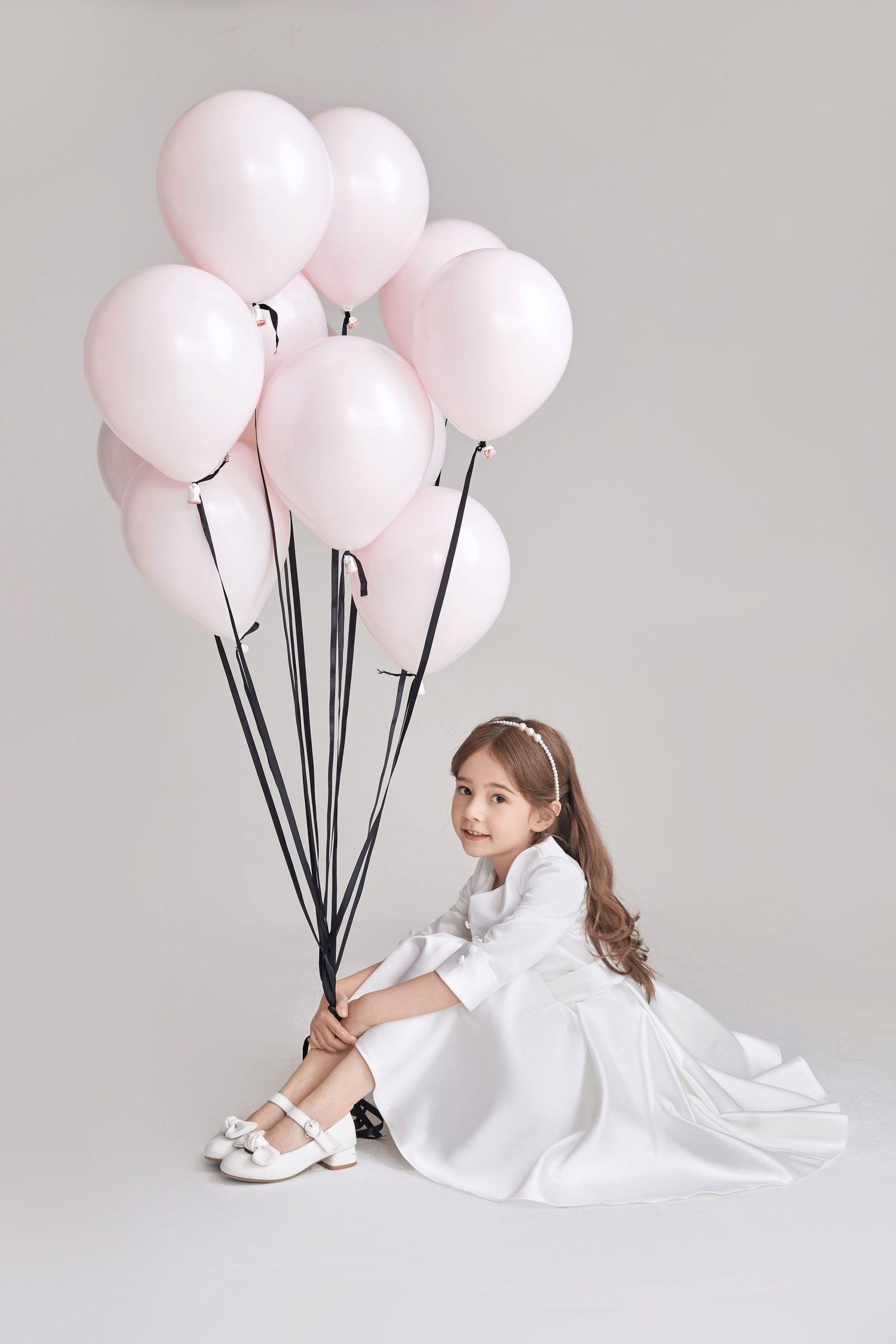 Young girl in white dress sits on floor holding pink and white balloons on black strings.