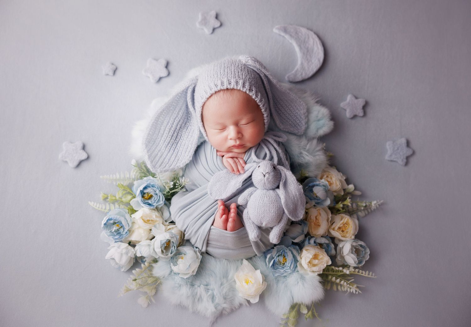 Newborn baby in gray bunny ear hat sleeping with knit rabbit toy surrounded by blue and white flowers.
