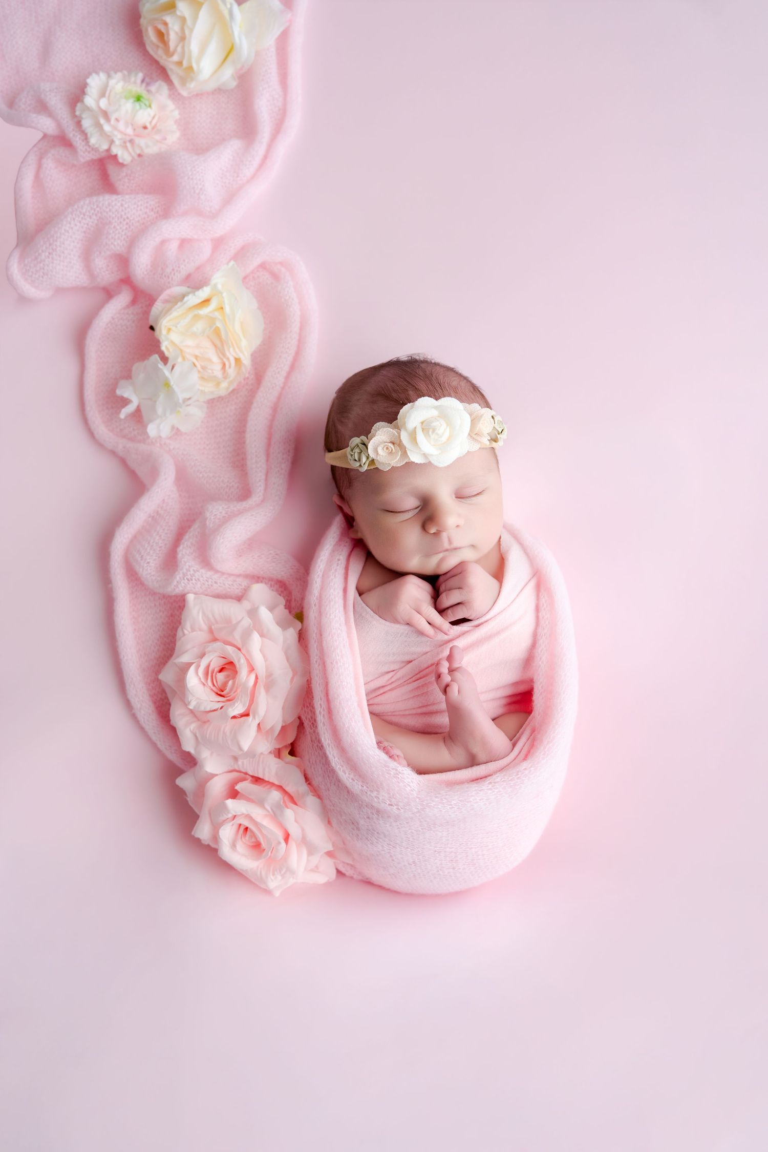 Sleeping newborn baby girl wrapped in pink swaddle with floral headband, surrounded by roses on pink background.