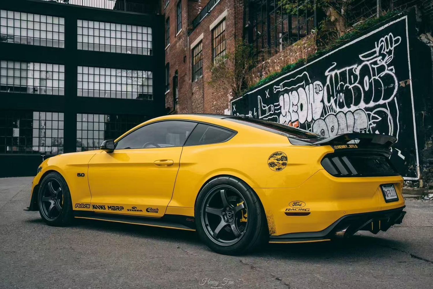 A bright yellow Ford Mustang GT with black rims parked in front of a graffiti-covered brick wall in an urban setting.