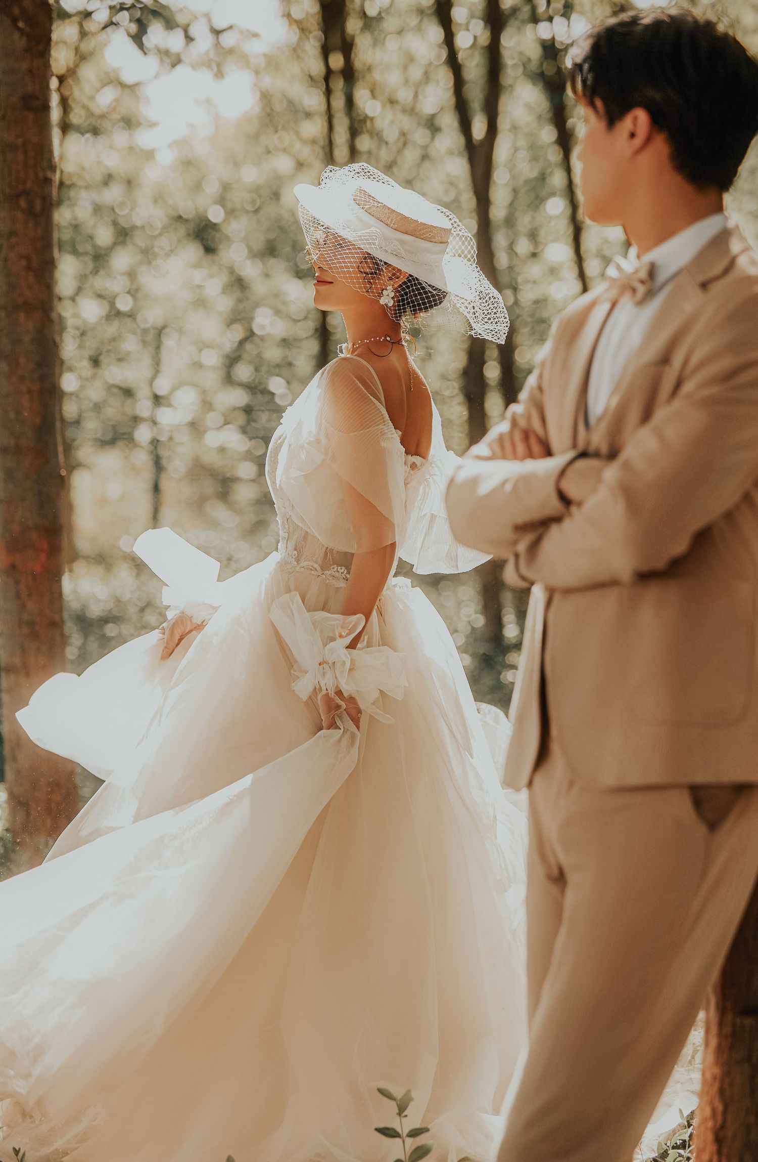 Bride in voluminous white tulle gown and veiled hat twirls in sunlit forest while groom in beige suit watches.