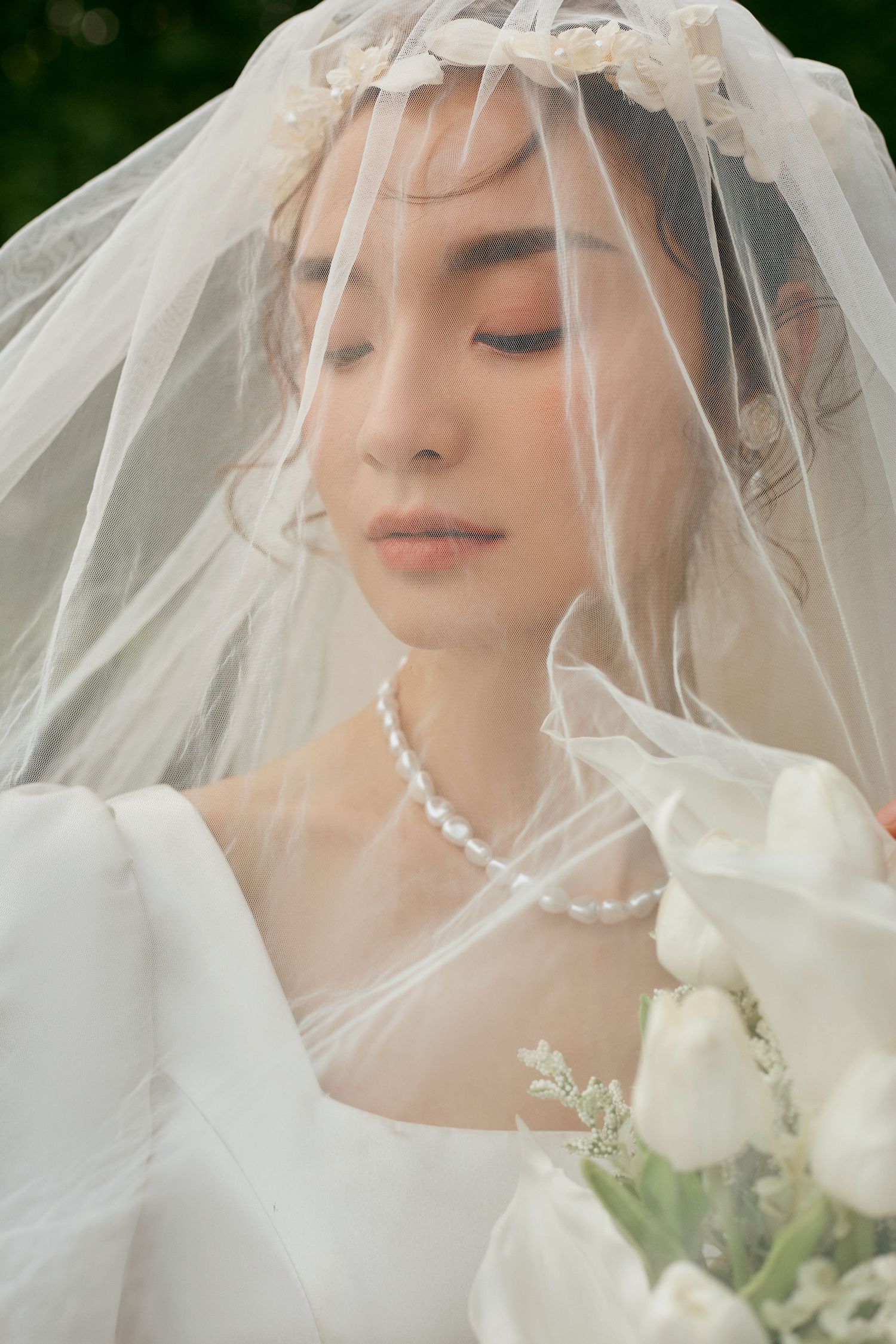 Serene bride with closed eyes wearing floral crown, pearl necklace, and veil holding white tulips.