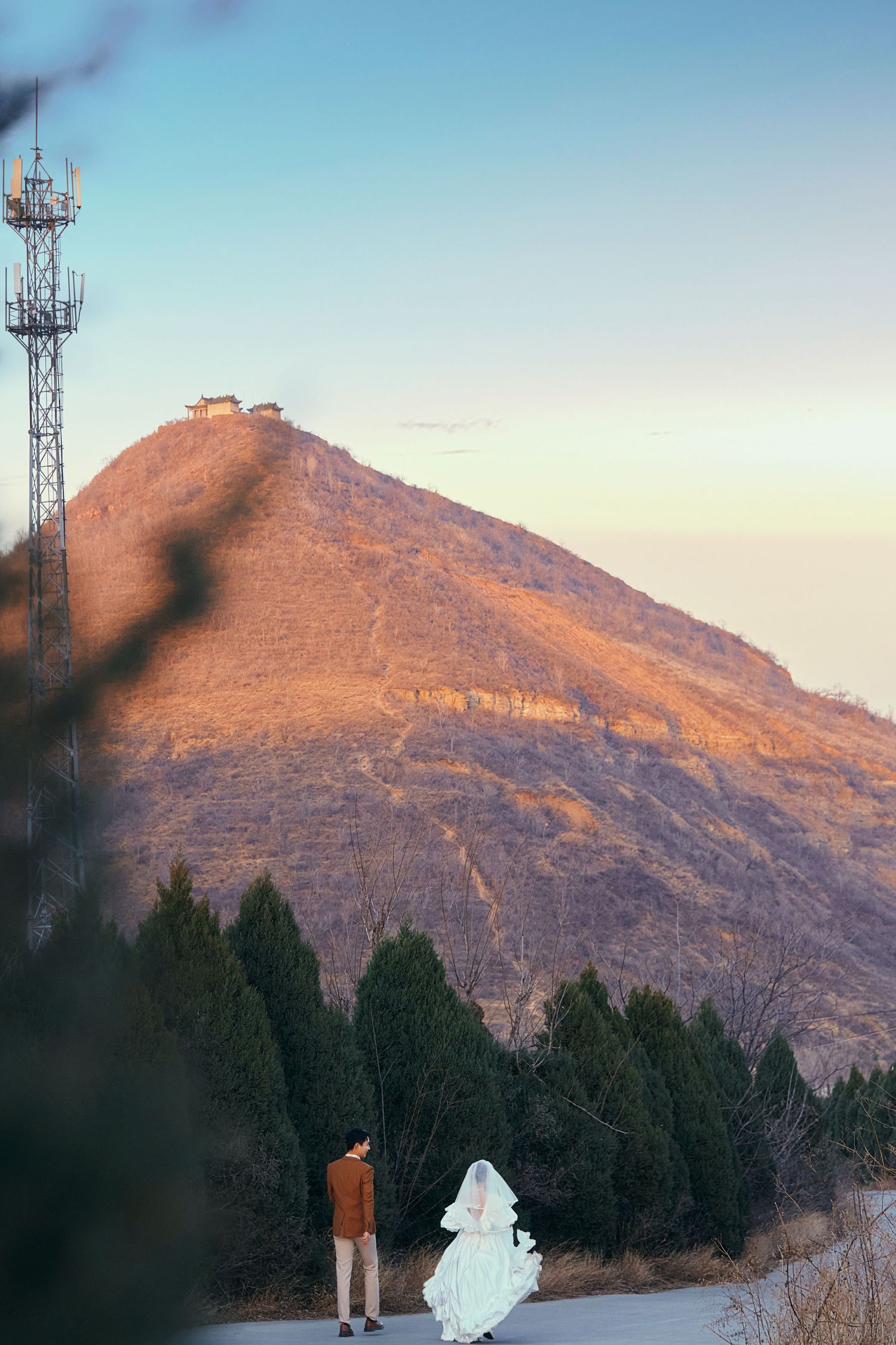Bride and groom standing before golden sunlit mountain at sunset during outdoor wedding photo session.