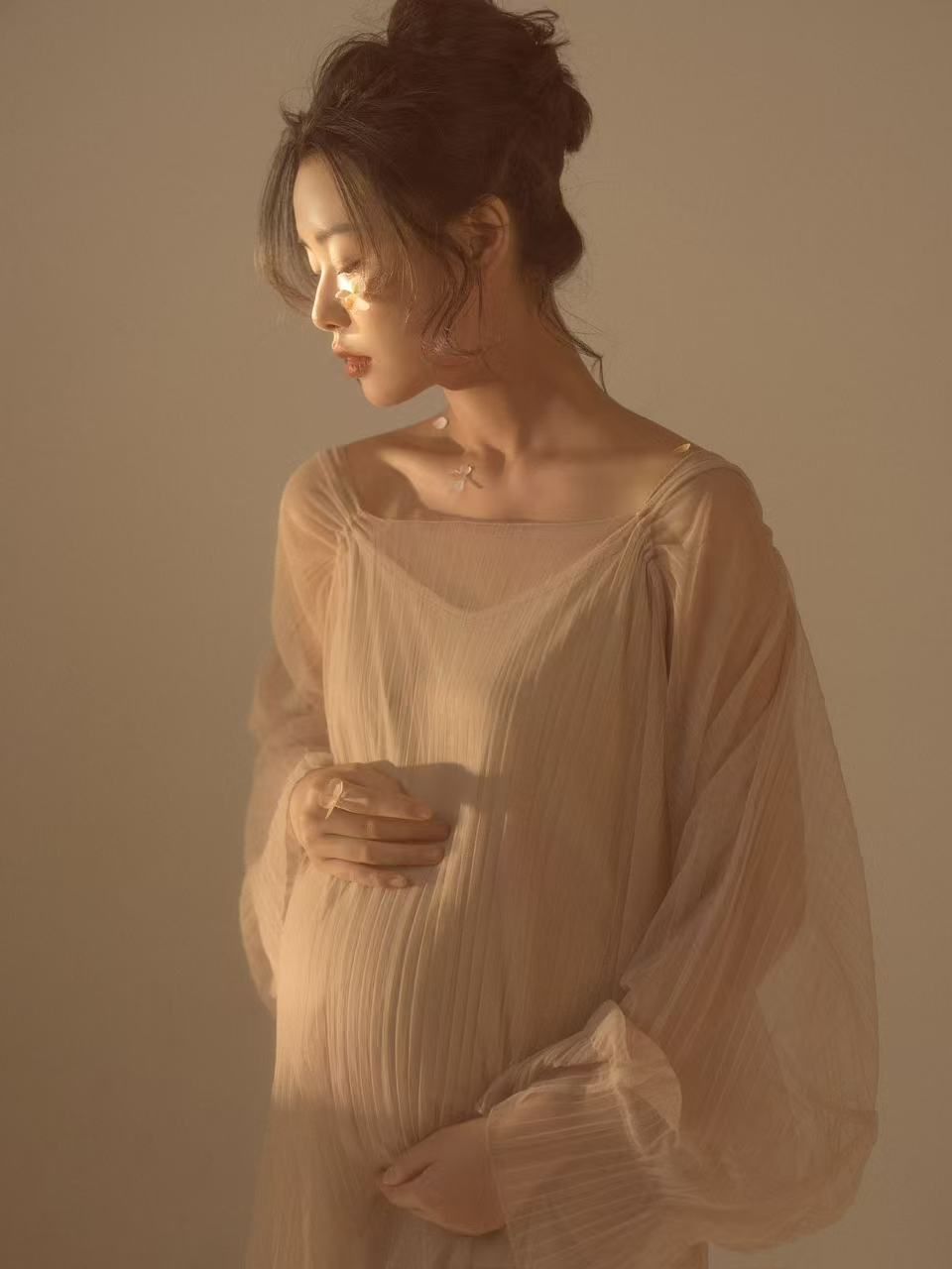 A pregnant woman in a sheer beige flowing dress cradles her baby bump in soft warm golden light.