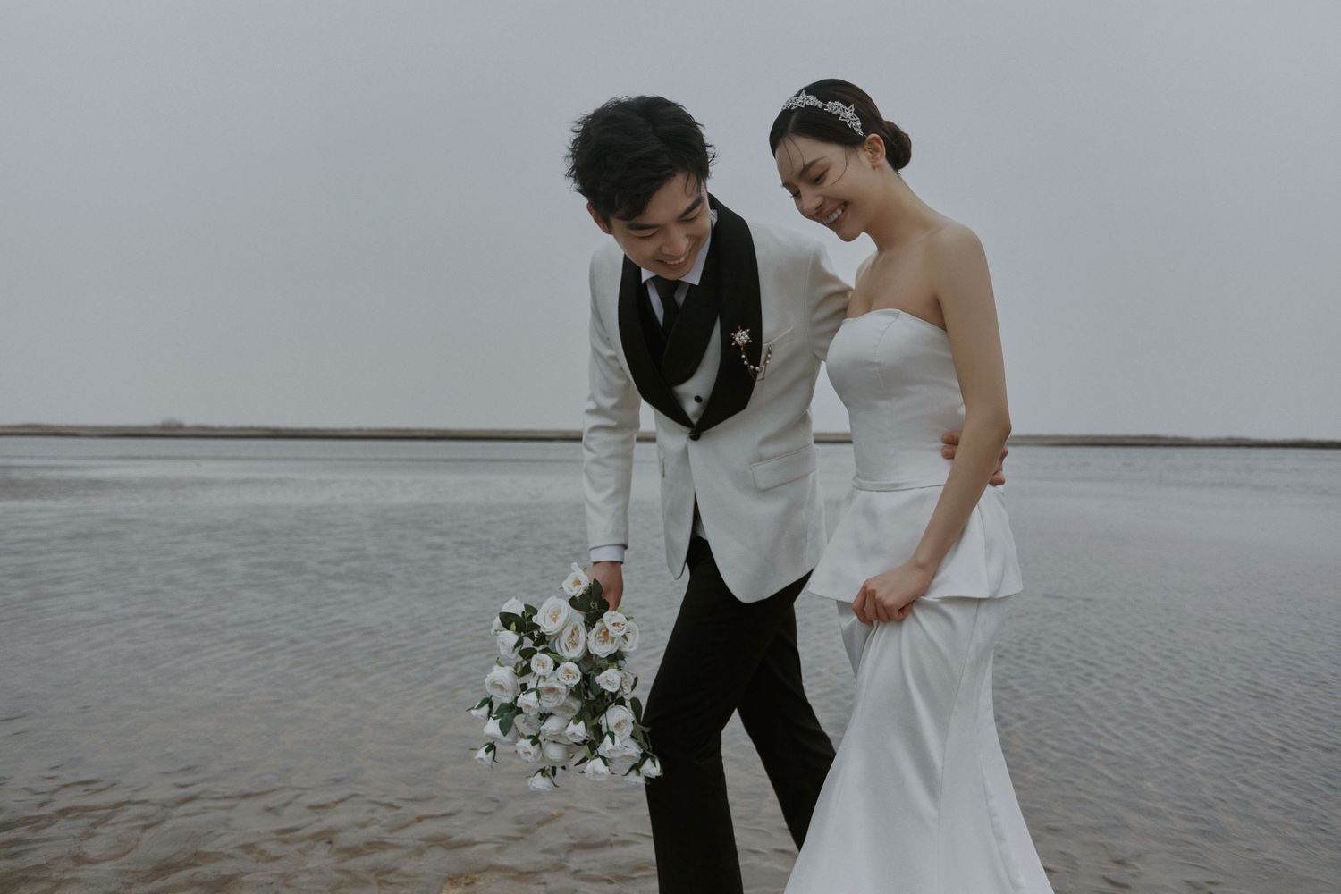 Bride in strapless mermaid gown and groom in white blazer walk along tranquil beach with white roses.