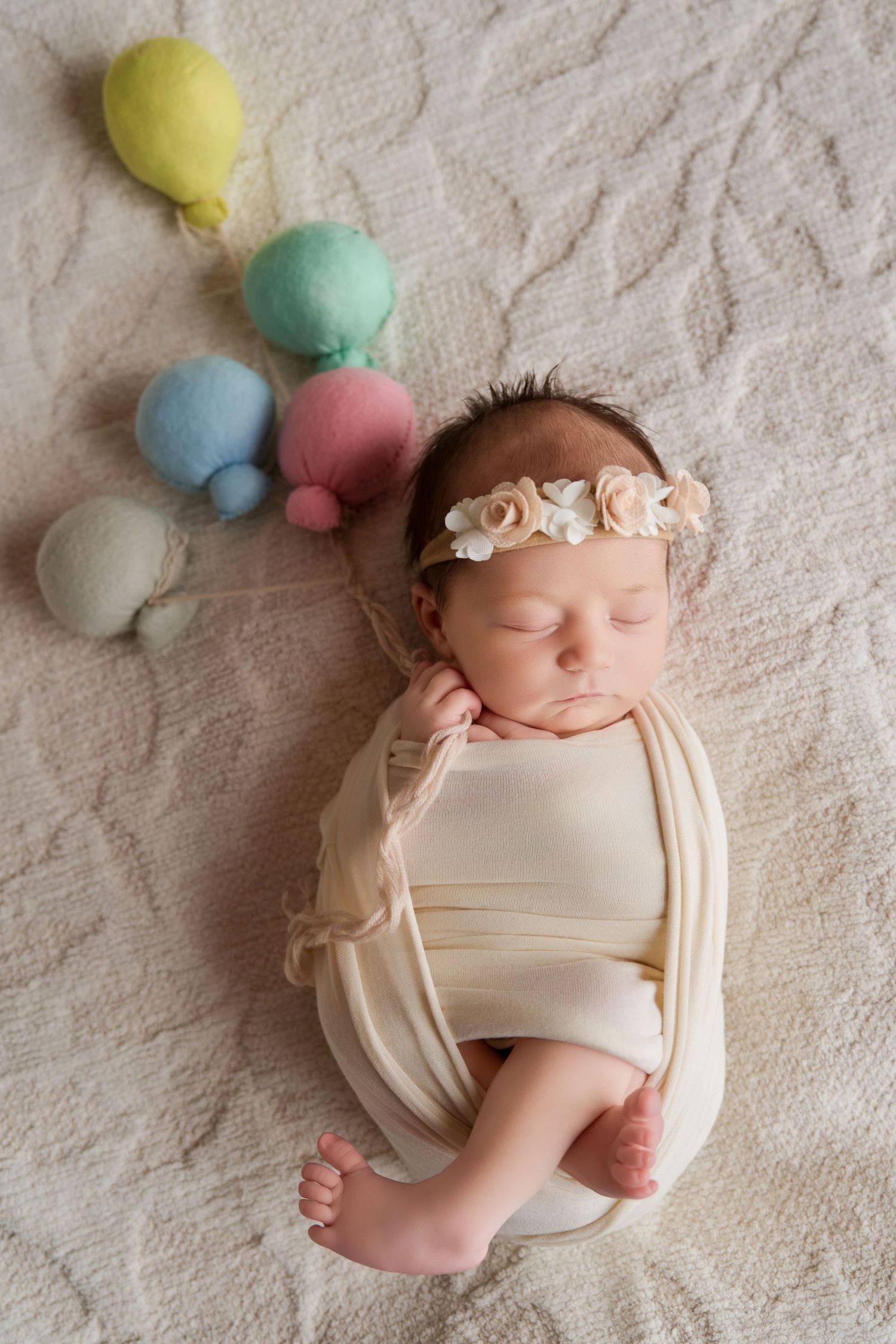 Newborn baby girl swaddled in cream wrap, wearing floral headband, sleeping peacefully beside colorful balloon props.