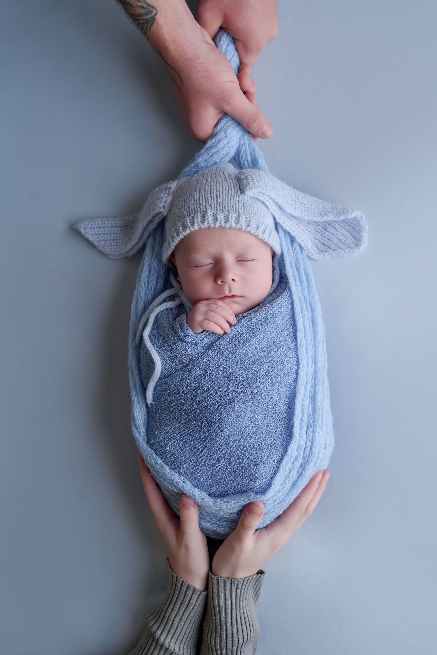 Newborn baby sleeping peacefully wrapped in blue knit blanket with bunny ear hat, cradled gently by parents hands.