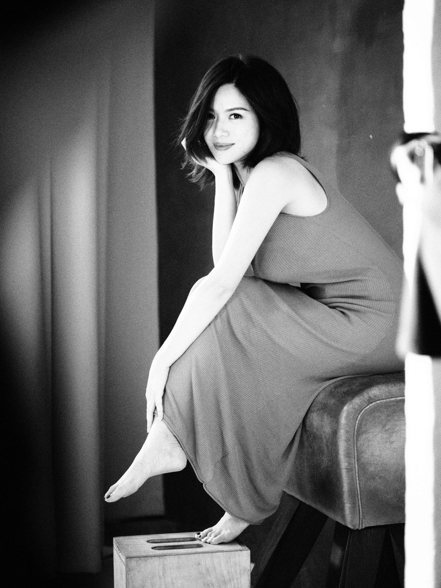 Black and white photo of a smiling woman in sleeveless dress sitting barefoot on a leather ottoman.