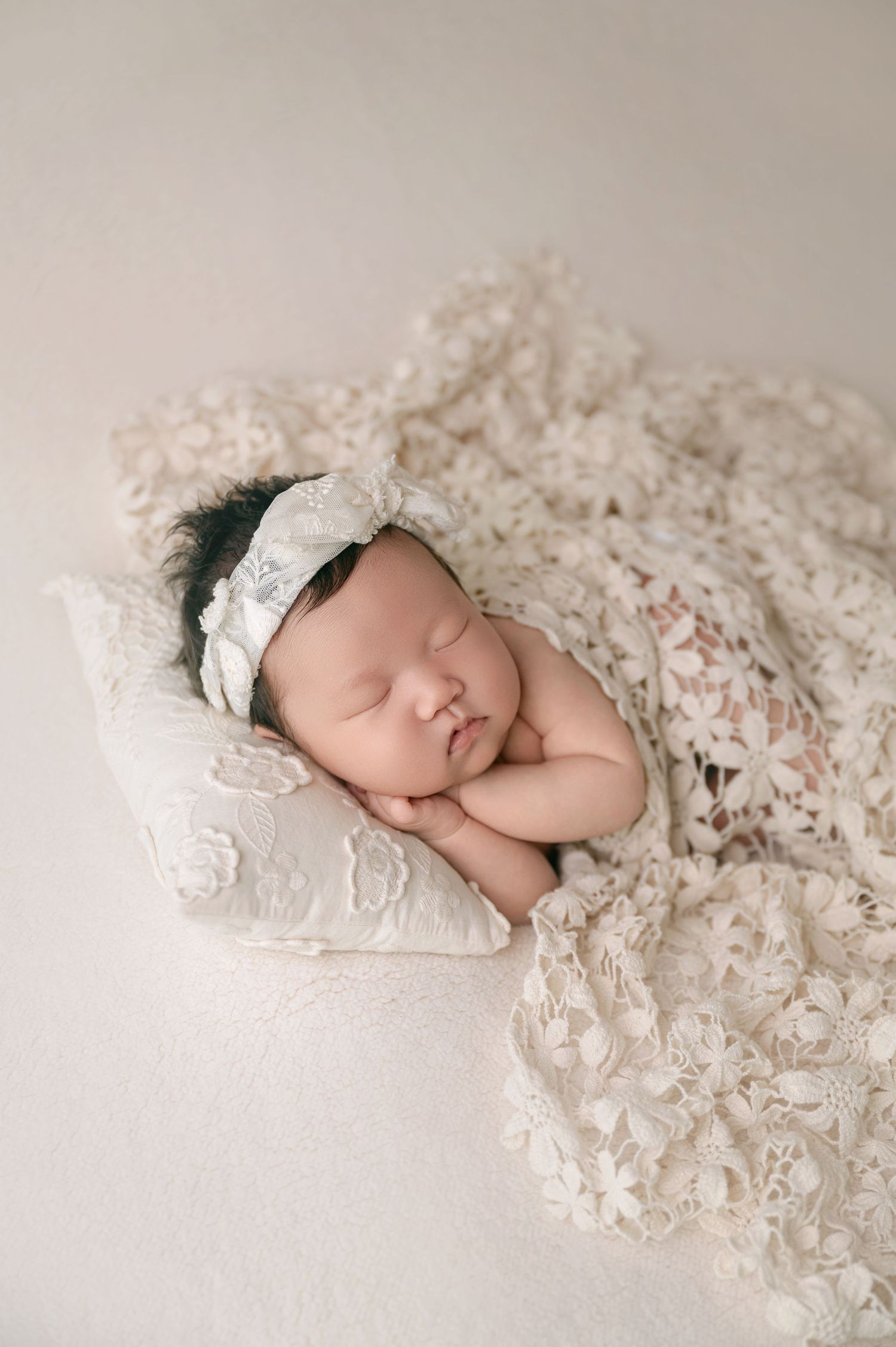 Peaceful newborn baby girl wrapped in cream lace blanket with floral headband.