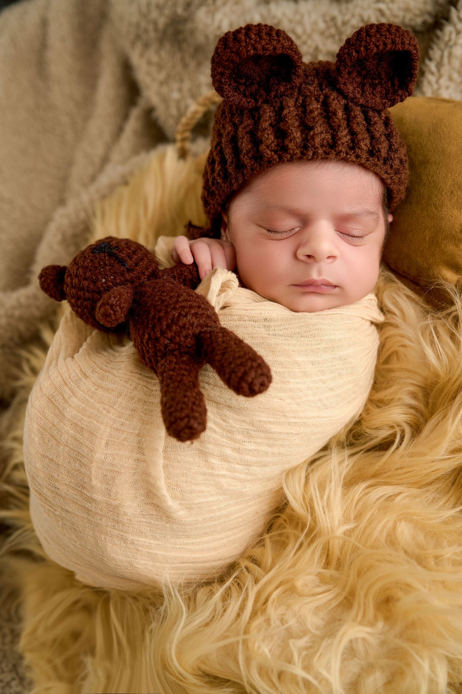 Sleeping newborn baby wrapped in cream swaddle, wearing brown knit bear ear hat, holding matching crocheted teddy bear.